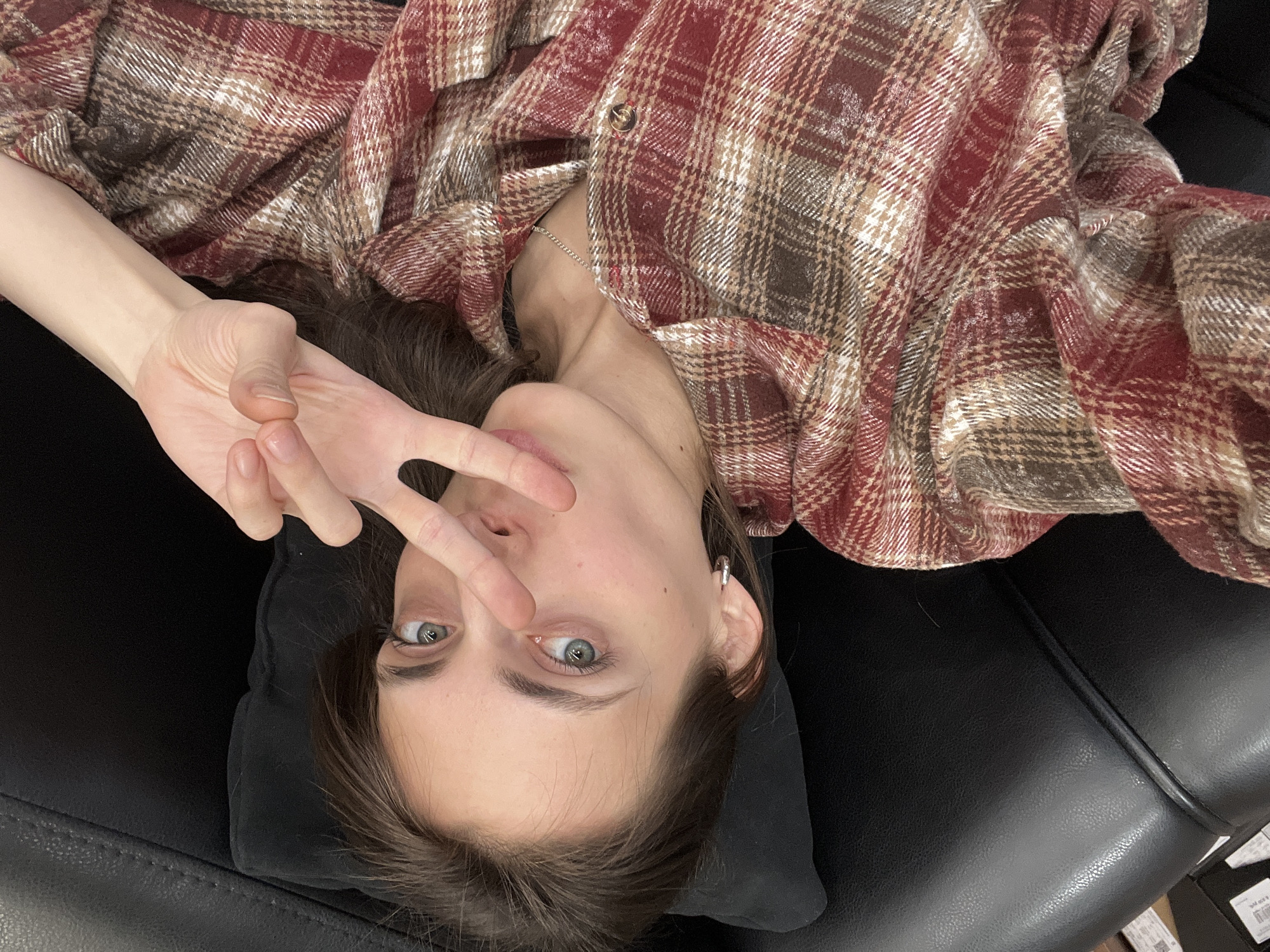 A young woman lies supine on a black leather surface, possibly a couch or chair, with a dark pillow beneath her head. She is wearing a red and brown plaid flannel shirt. Her right hand is raised, making a peace sign. Her face is centered in the frame, with her blue-grey eyes gazing towards the camera. The location appears to be an indoor setting. There are no other people or distinct objects visible to allow the determination of the city of Krasnoyarsk, Russia.