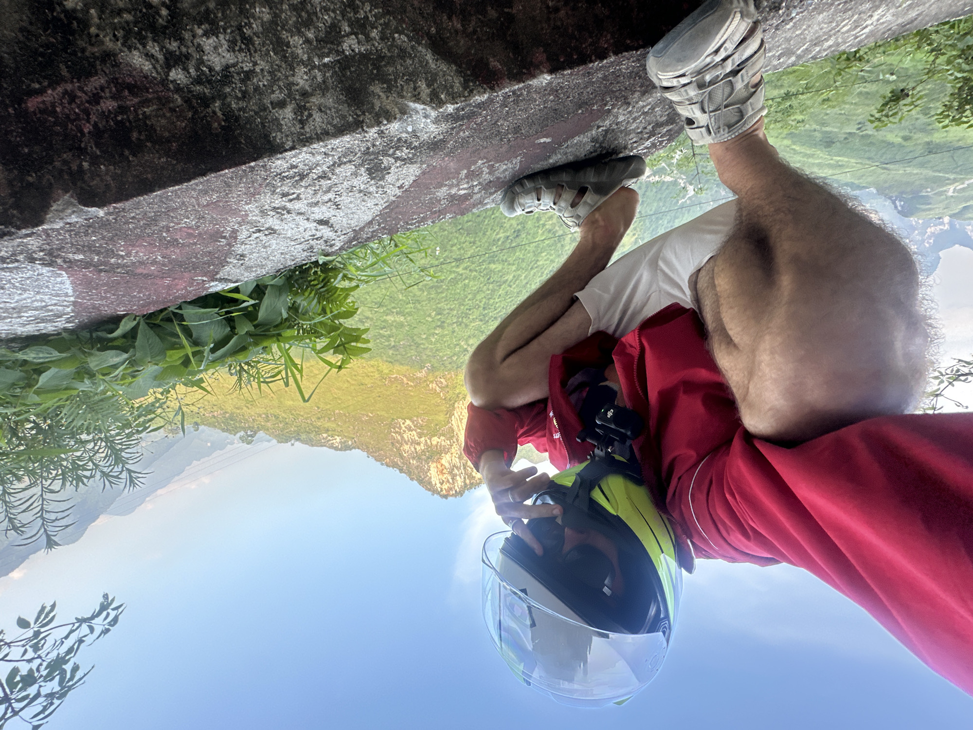 A person wearing a red jacket, white shirt, grey sandals, and a yellow and black helmet is photographed standing on the edge of a concrete structure in Đường Thượng Commune, Vietnam. The person is holding the helmet's visor with their right hand. A camera is mounted on the red jacket. Green plants are visible near the structure. A valley and mountainous landscape are in the background, with the sky visible in the lower part of the image.