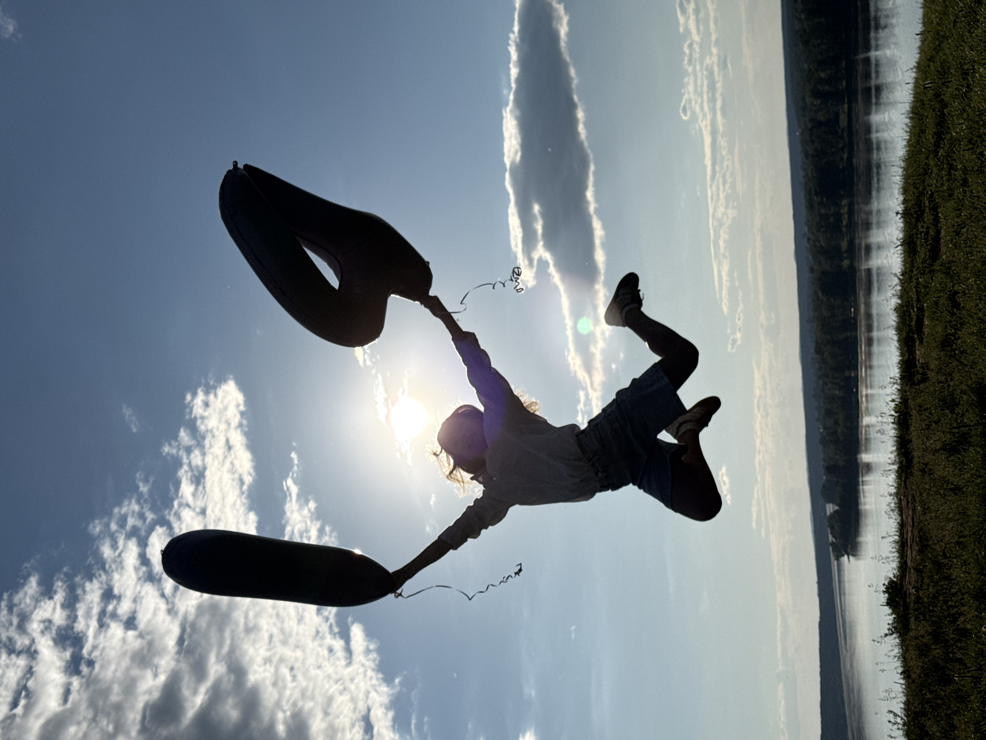 A lone individual is captured mid-jump, silhouetted against a brightly lit, partly cloudy sky. The subject, appearing to be a young female with hair flowing outwards from her head, is positioned centrally in the frame, with arms extended upwards and outwards. In each hand, she holds the string of a large, dark, number-shaped foil balloon. The balloon held in her left hand is elongated and cylindrical, resembling the number '1'. The balloon in her right hand is broader and curved, consistent with the shape of a '0', '6', or '9'.
The sun is directly behind the subject's head, high in the upper center-right sky, emitting a strong glare that creates a prominent lens flare and bright halo around its position. The sky is predominantly clear blue with scattered white cumulus clouds, some illuminated by the sunlight and others casting subtle shadows.
Below the horizon, a wide body of water, likely a river, stretches horizontally across the midground, reflecting shimmering vertical streaks of sunlight on its surface. The far bank of the river is densely lined with dark green foliage, indicative of forested land. The foreground consists of a gently sloping grassy embankment, covered in various low-lying green plants and blades of grass, partially visible beneath the subject's feet.
The scene is strongly backlit, rendering the subject and the balloons almost entirely in silhouette, while the surrounding environment remains partially illuminated. This outdoor daylight scene is located in Ольховка, Russia.