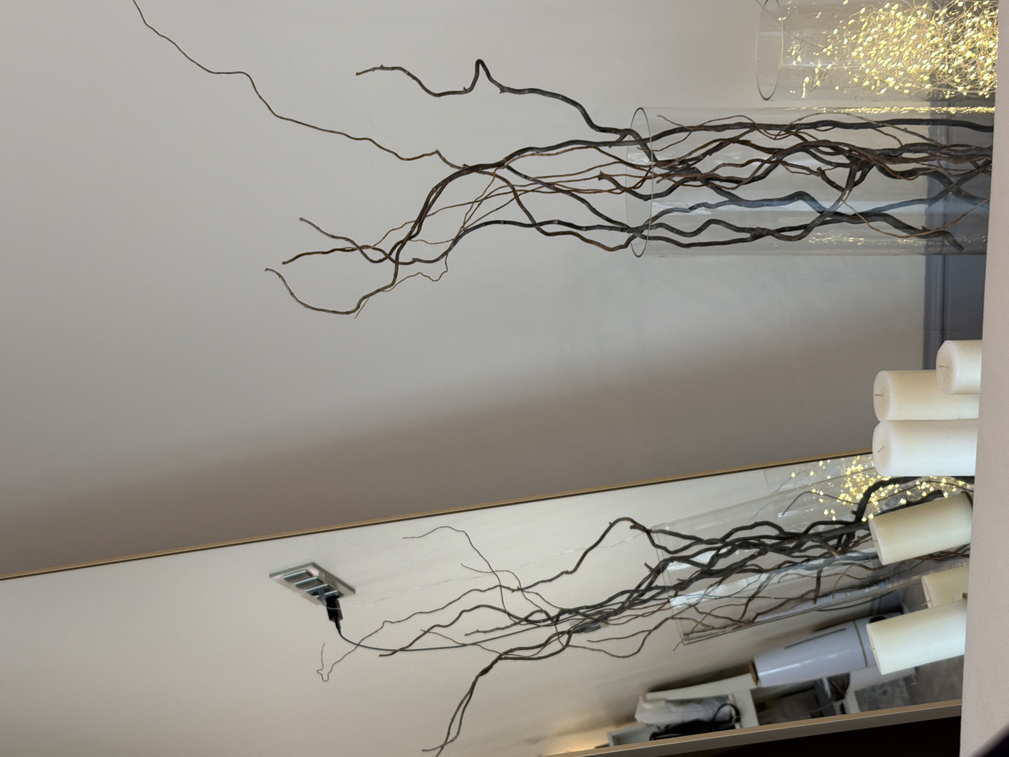 A room with a decorative display featuring bare branches arranged in a clear glass vase filled with string lights. The display is partially reflected in a mirror. Artificial candles are situated near the vase, and an electrical outlet is visible on the wall. Other household items can be seen in the reflection.