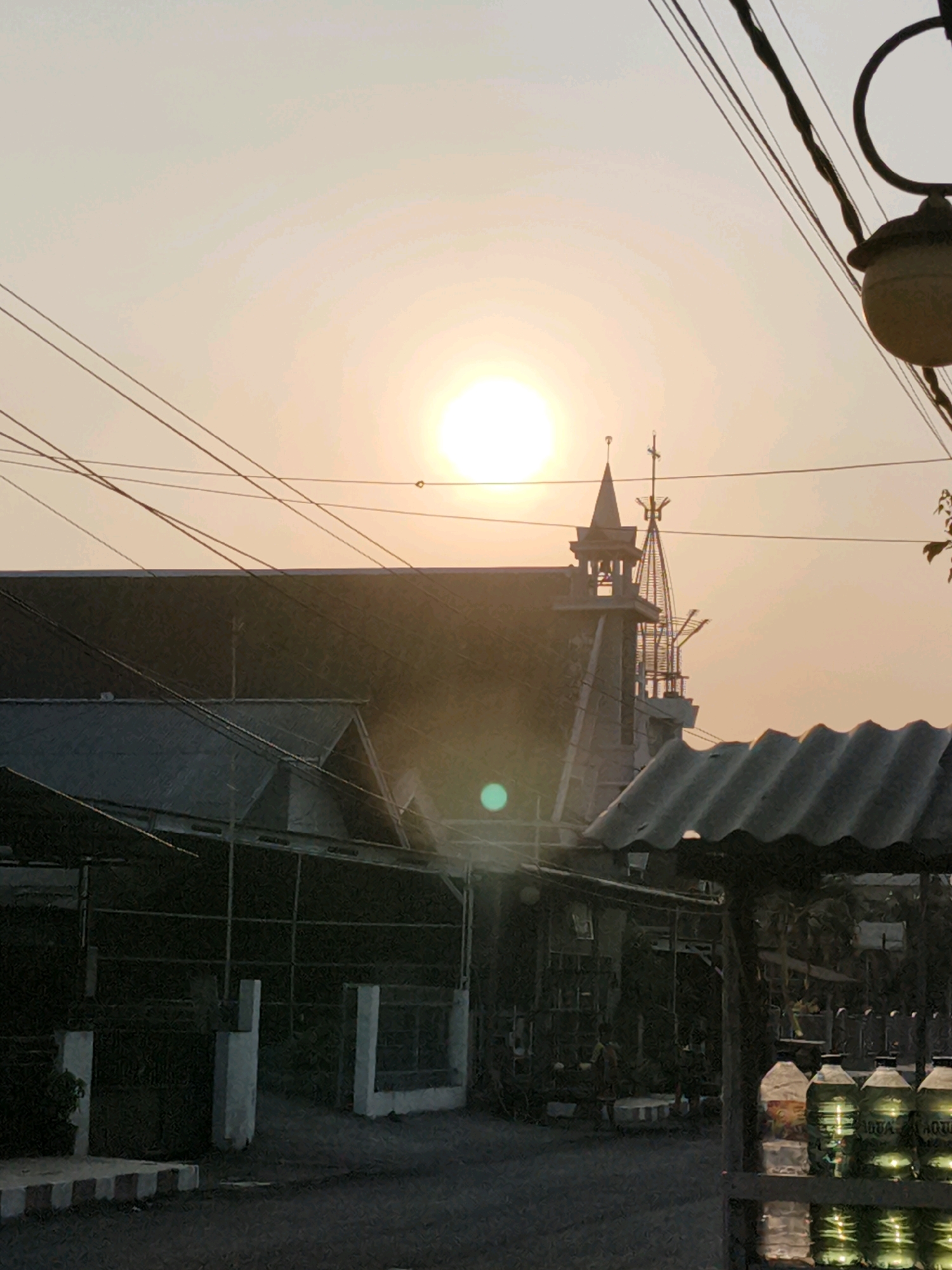 The image captures a sunset scene in Tombatu, Indonesia, with the sun positioned in the upper center of the frame. Overcast skies diffuse the sunlight, casting a soft, warm glow. A building with a steeple, possibly a church, is visible behind a series of utility wires that crisscross the sky. In the foreground, a corrugated metal awning shelters a display of bottled water, with the brand "AQUA" clearly legible on several bottles. The overall ambiance suggests a quiet evening in a semi-urban or rural setting within Tombatu.