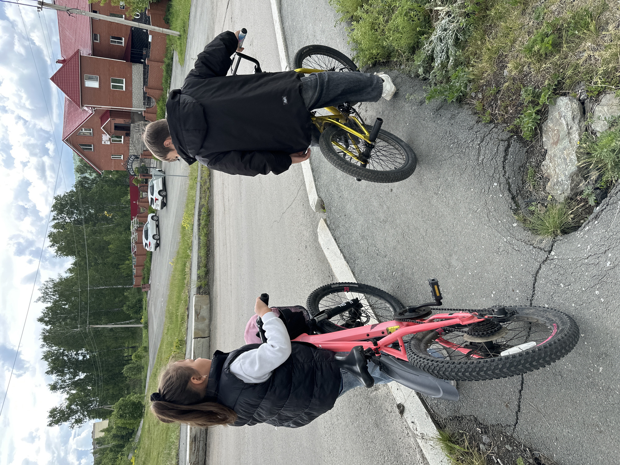 The image captures two young individuals, a boy and a girl, standing outdoors on a paved surface next to their bicycles. The photograph is oriented vertically, appearing to have been taken with the camera rotated.
In the upper right portion of the frame, a boy is seen from behind, wearing a black puffer jacket with the brand name "AVIR" subtly visible on the back, dark pants, and white sneakers. He is standing near a bright yellow BMX-style bicycle, looking down towards it. The bicycle features black tires and what appear to be stunt pegs on the front and rear axles.
In the lower left, a girl is depicted wearing a black puffer vest over a white long-sleeved top, blue jeans, and dark boots. Her long brown hair is pulled back in a ponytail with a black scrunchie. She is positioned next to a pink bicycle, which appears to be a mountain bike with prominent knobby black tires, a black rear fender, and disc brakes. The tire sidewall clearly shows the text "MAXXIS" and "REKON RACE". A small pink bag is attached to the handlebars of her bike.
The setting includes a paved road or path with a grassy and rocky shoulder. In the background, there are several red-roofed brick houses, characteristic of residential architecture. One house prominently displays a sign with the text "ДВОРОВАЯ". Beyond the houses, numerous green trees are visible against a cloudy sky, indicating daytime. White cars are parked along a street in the mid-ground, and power lines traverse the scene. The ground is asphalt with some cracks, particularly near the bikes. The overall atmosphere is casual and observational, with no strong emotions evident from the subjects.