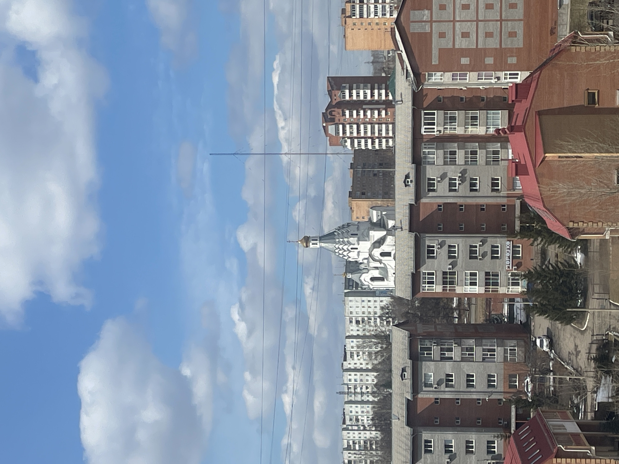 The image depicts a cityscape in Krasnoyarsk, Russia, under a partly cloudy sky. A prominent white cathedral with golden domes and intricate architectural details is centrally located. Residential buildings of varying heights and construction styles surround the cathedral, including structures with red and gray facades. Power lines crisscross the sky. The scene is captured from an elevated perspective. Trees are scattered among the buildings. A road with visible parked or moving cars is also seen.