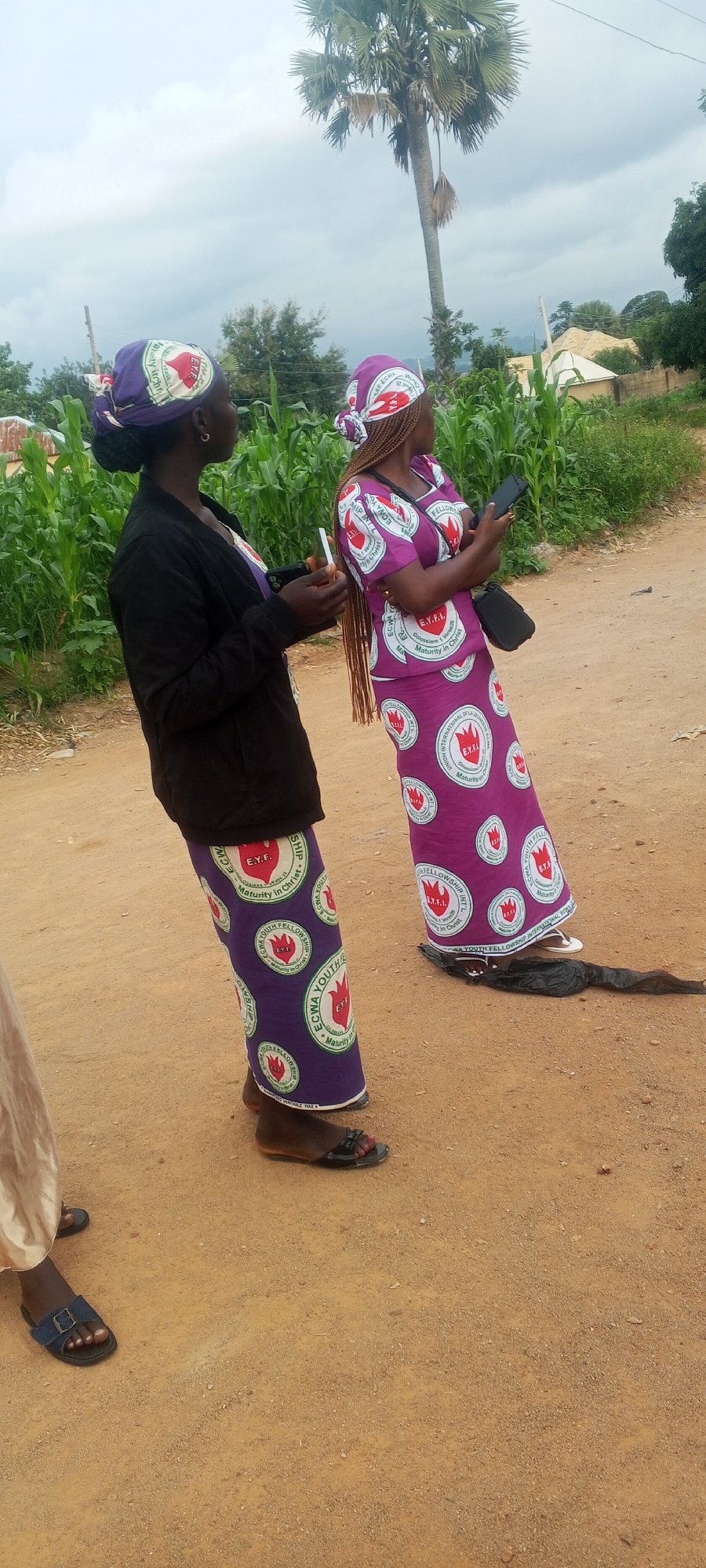 Two Nigerian women, identified as members of the ECWA Youth Fellowship, stand on a dirt road near a cornfield. The woman on the left wears a black jacket and a purple skirt with a repeating pattern of white circles containing a red flame logo and text. She holds a cigarette and a smartphone. The woman on the right wears a matching purple dress with the same pattern and has her hair braided. She is also holding a smartphone and has a black bag slung over her shoulder. A palm tree stands behind them, and the sky is overcast. The environment suggests a rural setting within the vicinity of Ohit, Nigeria, indicated by the typical Nigerian landscape and architecture visible in the background.