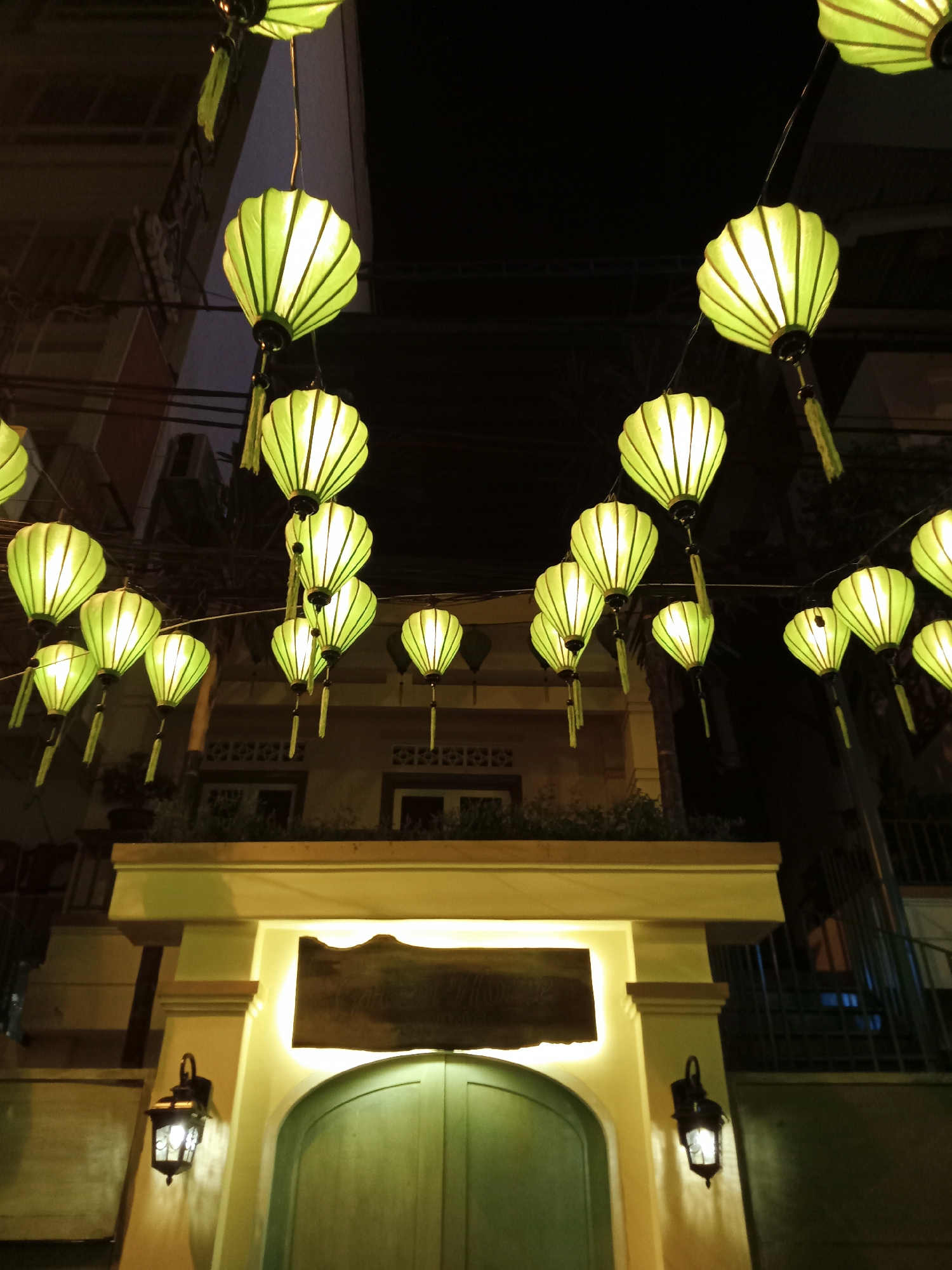 The image depicts a night scene in Nha Trang Ward, Vietnam, featuring rows of glowing green lanterns suspended above a building entrance. The lanterns are rounded with vertical lines, and each has a tassel hanging down. The building entrance is framed by a light yellow archway with a dark sign above a green arched door. Two lit wall-mounted lanterns flank the entrance. Dark buildings and street elements are visible in the background.
