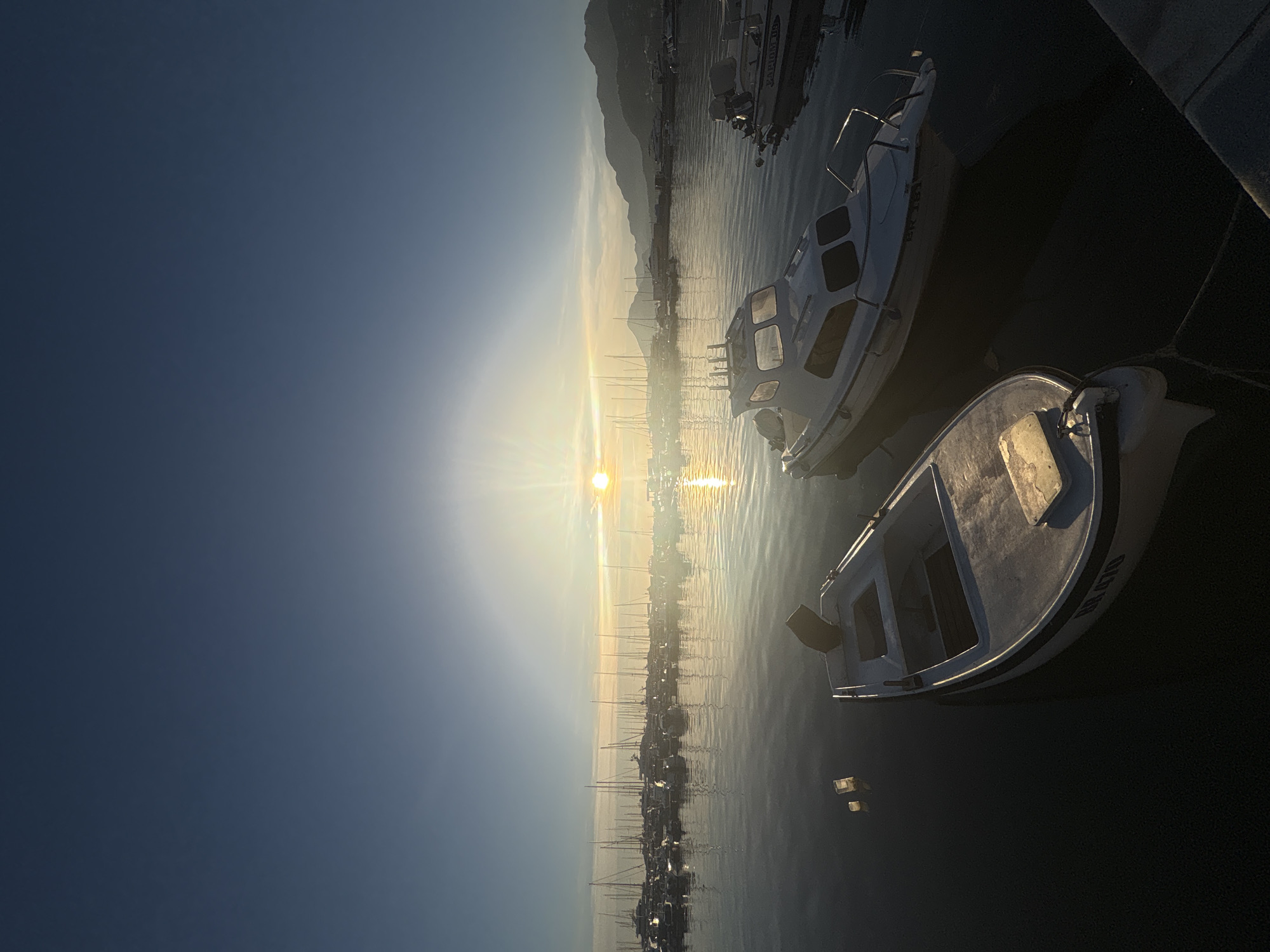 The image depicts a tranquil harbor scene at sunrise in the city of Bar, Montenegro. The sun, low on the horizon, casts a golden glow across the sky and water, creating a shimmering reflection. In the foreground, two white motorboats are docked. The boat on the left is smaller and appears to be a dinghy, while the boat on the right is larger with a cabin. The water is calm, with subtle ripples. In the background, a multitude of sailboats and yachts are moored, their masts creating a delicate silhouette against the brightening sky. Further in the distance, a range of hills or mountains forms the backdrop, partially illuminated by the morning sun. The overall atmosphere is one of peace and serenity.