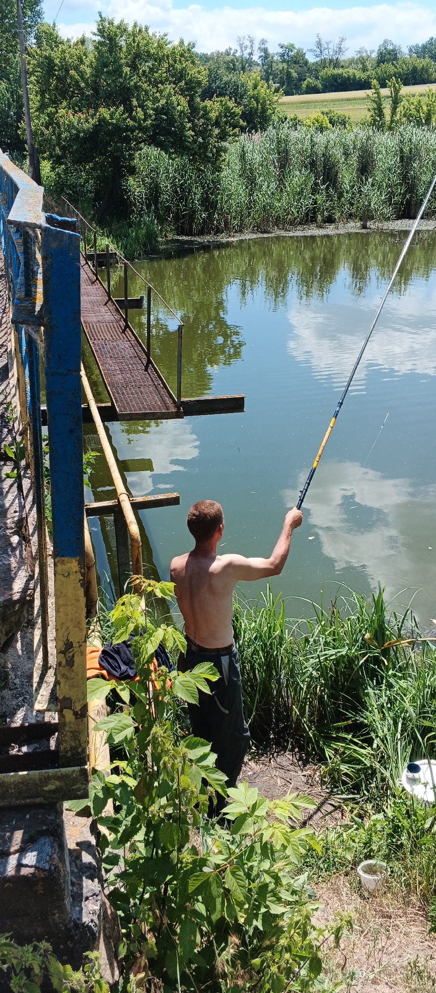 A shirtless man is fishing from the bank of a body of water, possibly a pond or small river, near a rusted metal walkway attached to a bridge. The man is facing away from the camera, holding a fishing rod that extends into the water. A fishing float is visible on the line. The surrounding environment is lush with green vegetation, including tall reeds and trees. The bridge structure is partially visible on the left side of the frame, painted blue and yellow. A pile of clothing, including an orange shirt and dark fabric, lies on the ground near the base of the bridge. The sky is partly cloudy. This scene is located in Ternivka, Ukraine.