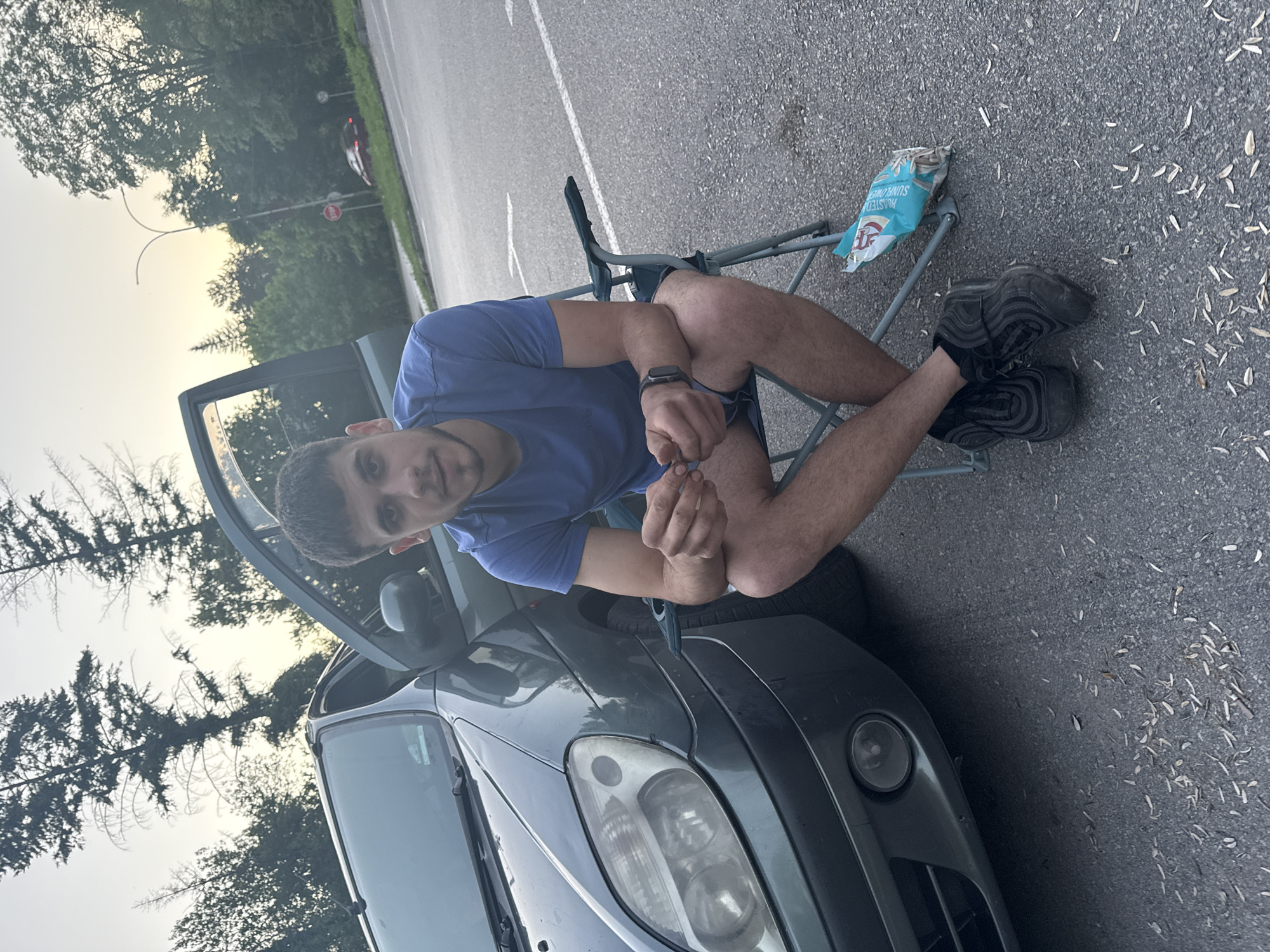 A young man is seated in a folding chair next to a gray car, its driver's side door ajar, on an asphalt surface. The location appears to be an outdoor setting, potentially a parking area or roadside in Pleven, Bulgaria, though specifics are not verifiable. He is wearing a blue t-shirt, dark shorts, and dark sneakers with black socks. He is holding something small in his hands. A discarded snack bag (sunflower seeds) rests against the chair. The area is littered with small, dry leaf debris. In the background, trees and foliage are visible, with a faint indication of street signage in the distance.