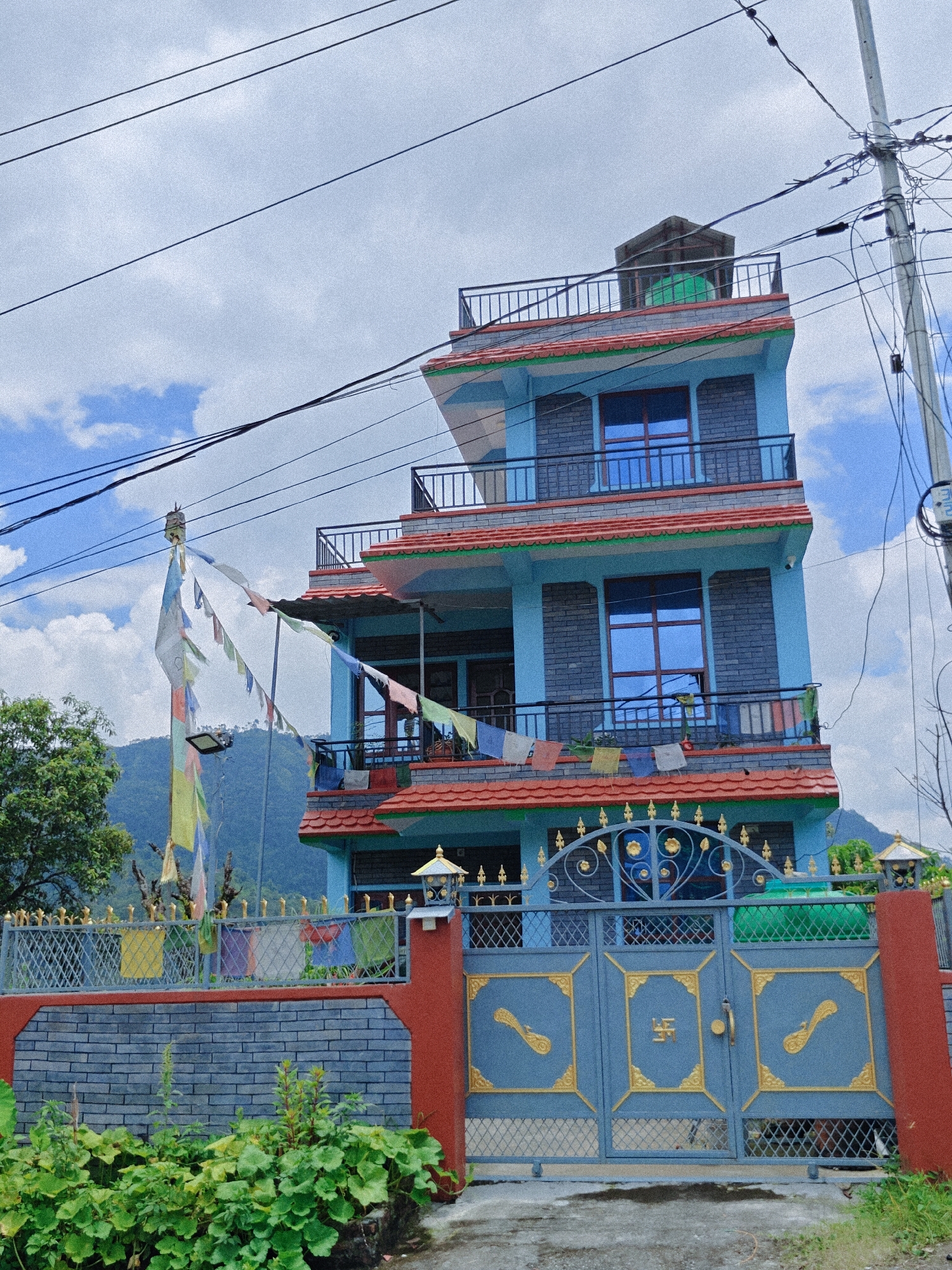 This image captures a multi-story building in Pokhara, Nepal, under a cloudy sky. The building is painted in shades of light blue with red tiled roofing and decorative green trim. It features multiple balconies with metal railings and dark grey brickwork on some sections. Prayer flags, colorful and fluttering in the wind, are strung from a pole on the left side of the building. A large, ornate blue metal gate with gold detailing, including a swastika symbol, stands at the entrance, bordered by a brick wall and lush green foliage. Utility poles and numerous electrical wires crisscross the sky above the building, indicating an urban or semi-urban setting. In the background, rolling hills or mountains are visible, partially obscured by mist. There are no people visible in the image.