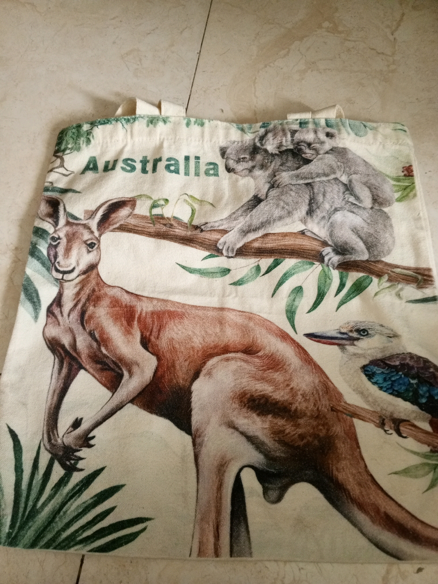 The image shows a close-up of a reusable tote bag featuring a printed design of Australian wildlife. The design includes a kangaroo, a mother koala with a joey on her back, and a kookaburra perched on a branch. The word "Australia" is printed in green text above the koalas. The tote bag is made of a light-colored fabric and has two white handles at the top. The bag is lying flat on a beige tiled surface. The scene is brightly lit, and the colors of the print are vibrant. There is no discernible connection to Masbate City, Philippines.