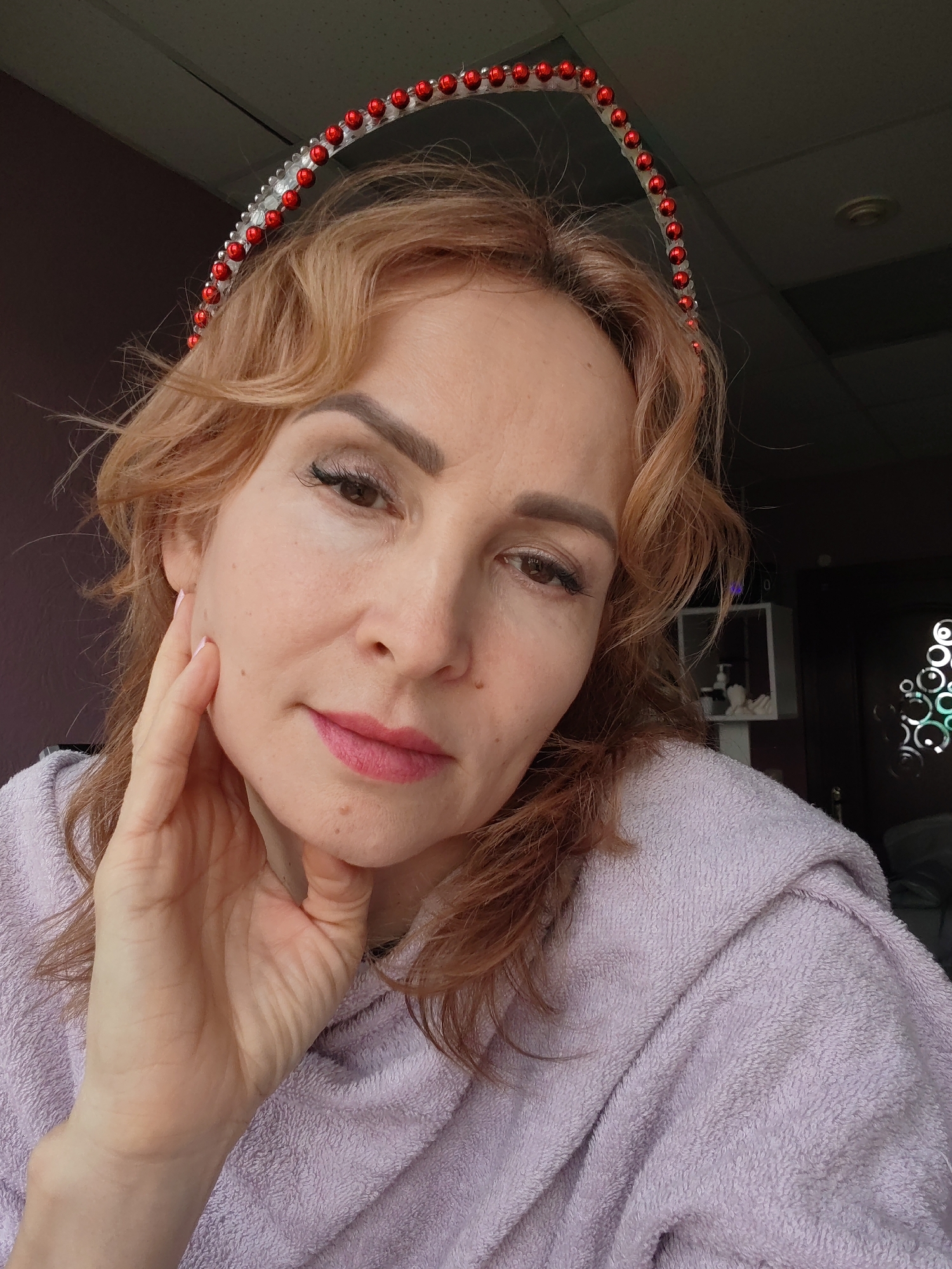 A woman with light reddish-blonde, wavy hair is photographed from the chest up, indoors. She is wearing a red and clear beaded headband. Her left hand is touching her cheek. She has fair skin, and is wearing light makeup, including pink lipstick and eyeliner. She appears to be wearing a hooded, light purple terry cloth robe or towel. The background is indistinct, showing a wall in a dark shade of purple or brown, part of a dark-colored door with decorative cutouts, and a white shelf with bottles. The location is Chaikovsky, Russia.