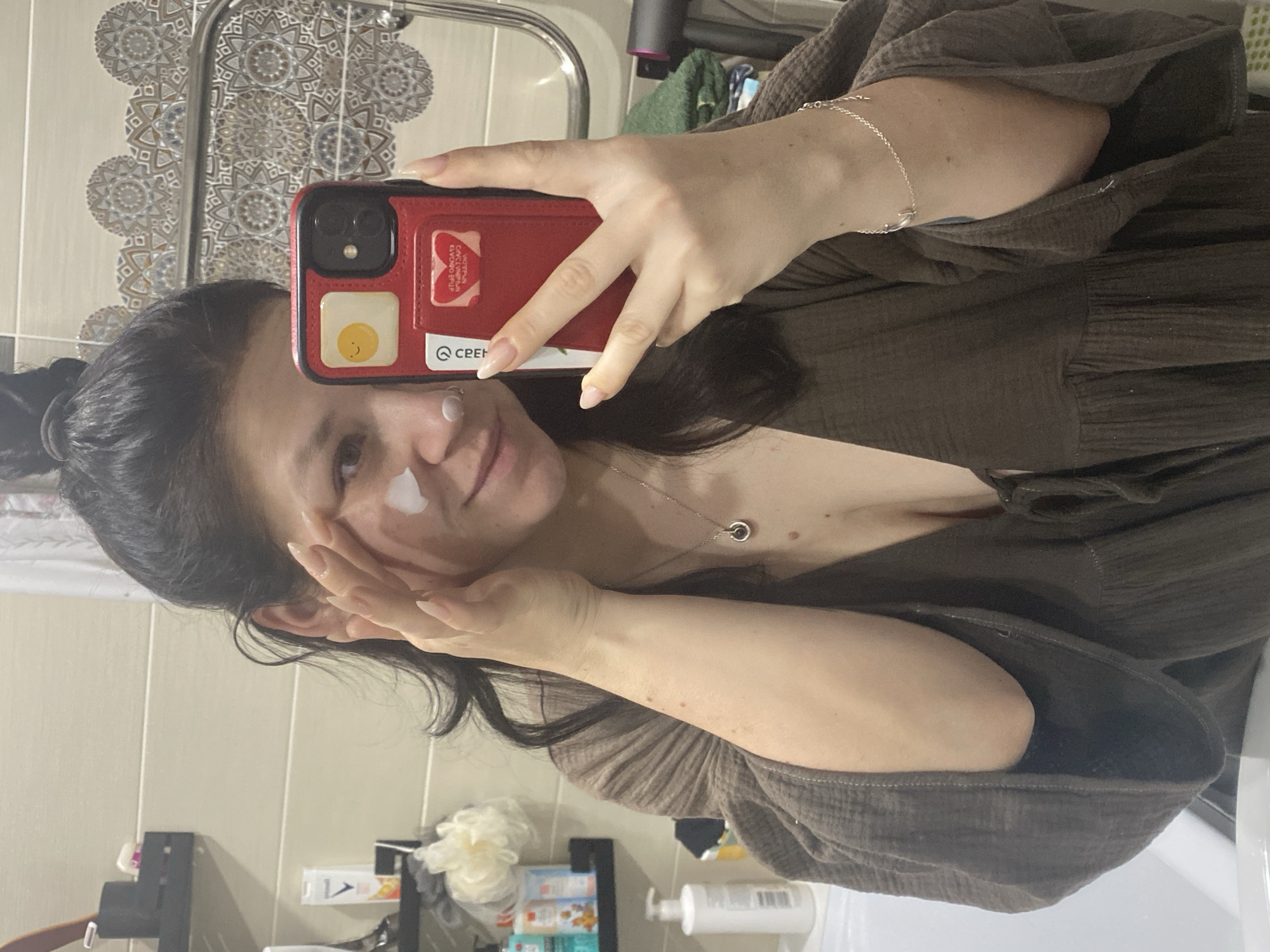 A woman is taking a selfie in a bathroom. She is holding a red phone with a cartoon fried egg on the back. She has a small dollop of white cream on her left cheek and is applying more with her right hand. She is wearing a brown top and a silver necklace. The bathroom has white tiles and a patterned border above the sink. Below the sink is a shelf with various toiletries. The location is in a bathroom in Chaikovsky, Russia.