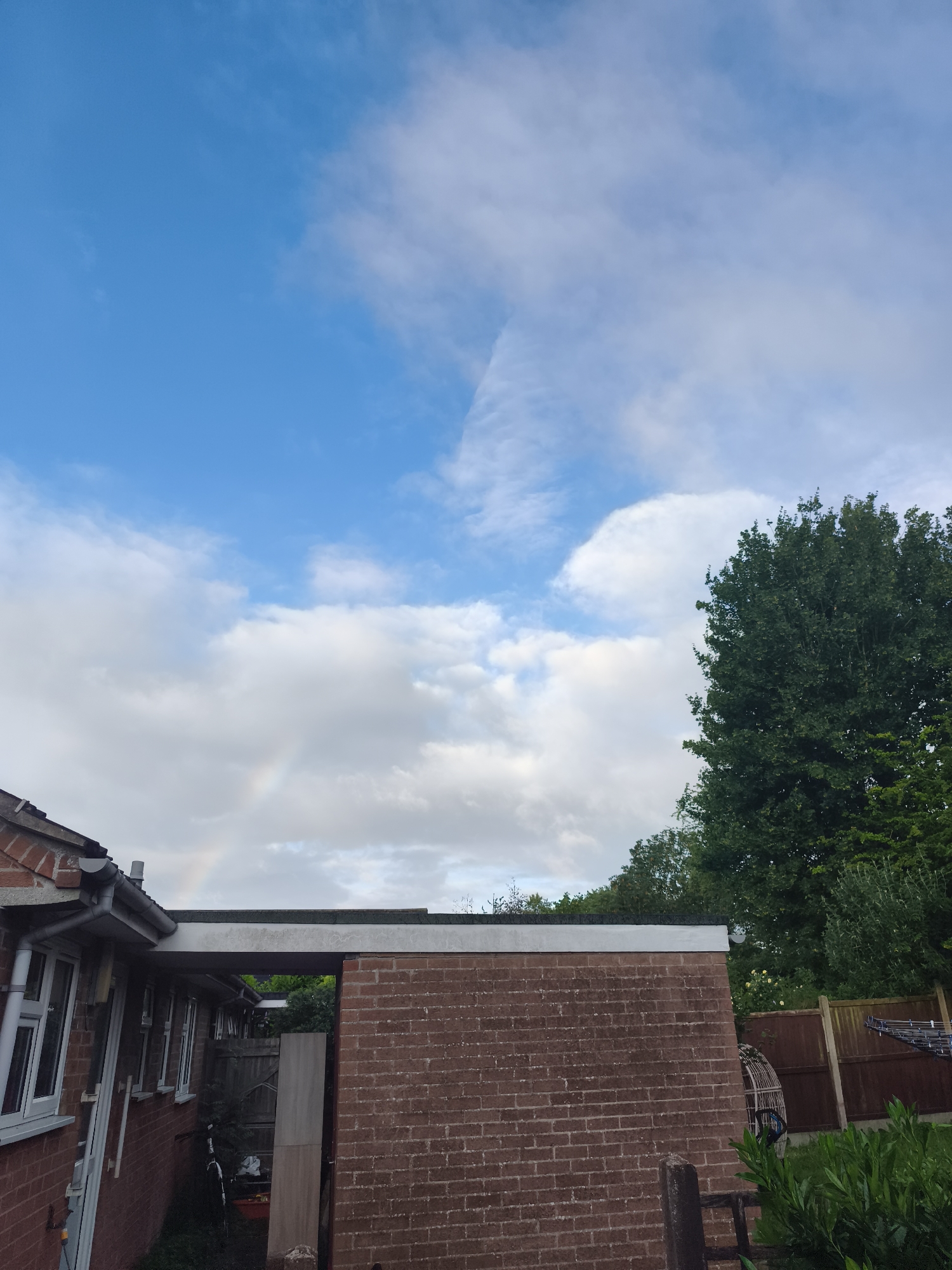 This image depicts a residential outdoor scene, likely a backyard or garden, under a sky with scattered white clouds and patches of bright blue. On the left, a section of a brick building with white-framed windows and a grey gutter system is visible. A prominent brick structure, possibly a garage or extension with a flat, dark grey roof, occupies the midground on the right. Behind this structure, a wooden fence and a clothes airer can be discerned. A large, verdant tree stands tall on the far right, with other greenery, including bushes in the foreground, contributing to the natural elements of the scene. A narrow passageway between the buildings reveals a bicycle.
A notable detail is the faint, pastel rainbow arching subtly in the sky towards the lower-middle left, partially obscured by clouds, suggesting recent rain or atmospheric conditions conducive to its formation. The time of day appears to be daytime, possibly late afternoon, given the light and the presence of the rainbow. The sky also features an intriguing linear division in the clouds towards the upper center. No people or discernible activities are present, lending a quiet atmosphere to the residential setting. No text is visible in the image. The architecture and garden elements are consistent with a location such as Huntingdonshire, United Kingdom.