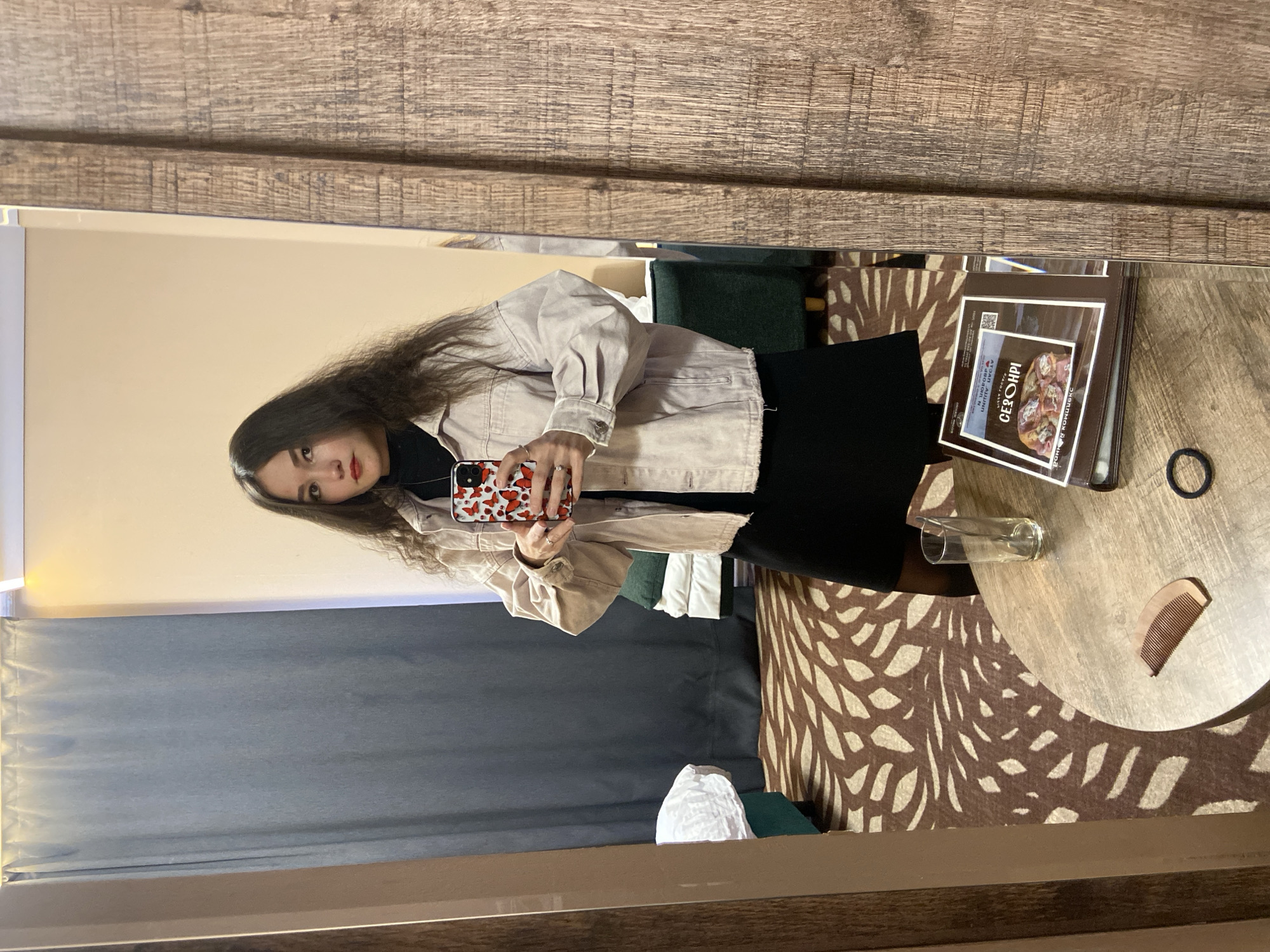 A young woman is captured in a mirror selfie, standing in what appears to be a hotel room or apartment. The image is taken from a slightly low angle. She has long, dark, wavy hair and is wearing a light-colored, possibly beige, denim jacket over a black turtleneck and a black skirt or dress. Her makeup includes red lipstick, and she has a neutral expression while looking directly into the camera. She holds a smartphone with a distinct red and white butterfly-patterned case in her right hand, taking the photo. A silver ring is visible on her left ring finger.
The room's interior is reflected in the mirror. To the left, dark gray curtains are visible, with a light-colored wall above them. The mirror itself is framed by a wooden border with a visible grain. On the right side of the reflection, a round, light-toned wooden table is present. Scattered on the table are a clear empty drinking glass, a small wooden comb, and a black hair tie. A stack of brochures or books is also on the table, featuring Cyrillic text. The most prominent word readable is "СЕЗОНРИ" (SEZONRI), with other smaller text like "АУСАН ЛИШКАН" and "ЛЮБОВ И ЖИЗНЬ" (Love and Life) partially visible, along with a QR code. A brown and beige patterned rug covers the floor area visible in the reflection, and a dark green upholstered piece of furniture is also partially seen. The room appears to be lit by indoor artificial light, suggesting a general indoor setting in Peresvet, Russia, as indicated by the Cyrillic text.