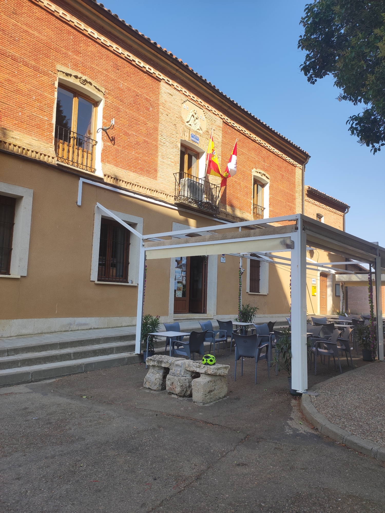 The image depicts the exterior of a building in Autillo de Campos, Spain, under a clear blue sky. The building is a two-story structure with a red brick upper facade and a tan stucco lower facade. The upper facade features several windows with wooden frames and decorative railings. Two flags, the Spanish flag and another red and white flag, are displayed from a balcony on the upper level. The lower facade has large windows with dark frames and a set of stone steps leading up to the entrance. In front of the building, there is an outdoor seating area with white tables and chairs covered by a retractable awning. A stone bench with some graffiti and a lime-green helmet rests on the pavement in front of the seating area. Trees are visible on the right side of the image. The scene appears to be a public building, possibly a town hall or a restaurant with outdoor seating.