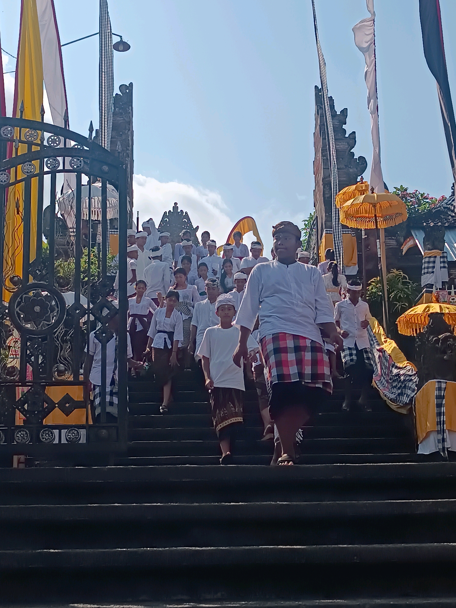 A group of people, mostly young males and females dressed in traditional Balinese attire, descend a wide stone staircase leading up to a temple. The attire includes white shirts and pants for some, while others wear white tops with sarongs or checkered cloths wrapped around their waists, and some have head coverings. The staircase is flanked by ornate gates on the left and temple architecture on the right, adorned with yellow banners and umbrellas. Sunlight illuminates the scene, casting shadows on the stairs. The temple structure appears to be made of stone and brick, with intricate carvings visible in the background. The atmosphere suggests a procession or a religious ceremony. This scene likely takes place at a temple in Tabanan, Bali, Indonesia, known for its rich cultural heritage and numerous temples.