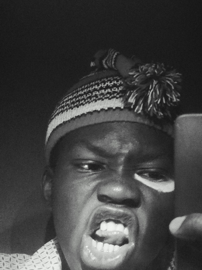A monochrome selfie captures a person wearing a beanie with a pom-pom. They are close to the camera, their face contorted in an intense grimace, exposing teeth and tongue. One eye is visible, while the other is partially obscured by the reflection of a light source. A cell phone is visible on the right, held by the person taking the photo. The background is dark and blurred.
