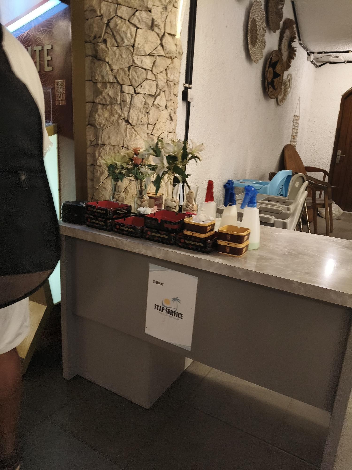The image shows a counter in what appears to be a restaurant or hotel lobby in Desa Wonoharjo, Indonesia. On the counter are several red and black ashtrays, some stacked and some with what looks like white paper or napkins inside. To the right of the ashtrays are several bottles, including white spray bottles and a red-capped bottle, some appearing to contain cleaning solutions. Behind the counter, a vase with white flowers is visible. A sign reading "STAFF SERVICE" with a palm tree logo is affixed to the front of the counter. In the background, a stone-clad pillar stands to the left, and a wall decorated with woven baskets is on the right. A stack of grey plastic chairs and a wooden chair are also visible in the background, along with a wooden door. A person's torso, wearing a black vest over a white garment, is partially visible on the far left of the image. The lighting suggests an indoor environment, possibly with artificial light sources.