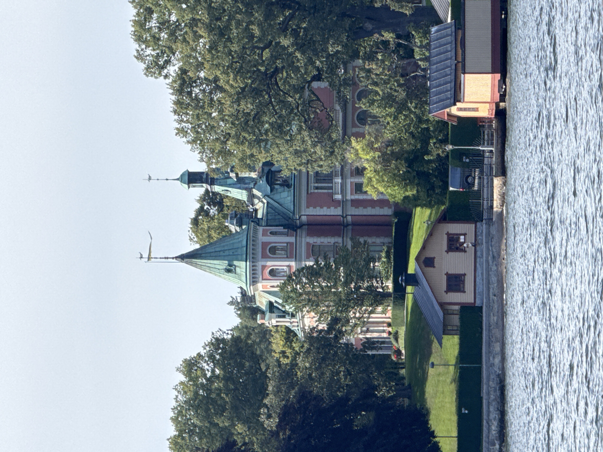 This image captures a view of a large, pink and green historic mansion situated behind lush green trees, with a body of water in the foreground. The mansion features distinctive pointed towers with copper or verdigris-colored roofs, arched windows, and decorative white trim. In front of the mansion, closer to the water, are two smaller wooden structures. The one on the left is a single-story building with a dark roof and yellow walls, featuring two windows and a small person walking past its right side. The structure on the right appears to be a boathouse or similar building with a similar dark roof and yellow and maroon colored walls. The water is choppy, suggesting a breezy day. The overall scene evokes a sense of historical grandeur and a peaceful waterfront setting. This appears to be Strandvägen in Stockholm, Sweden, a picturesque waterfront promenade lined with grand buildings and historic villas.