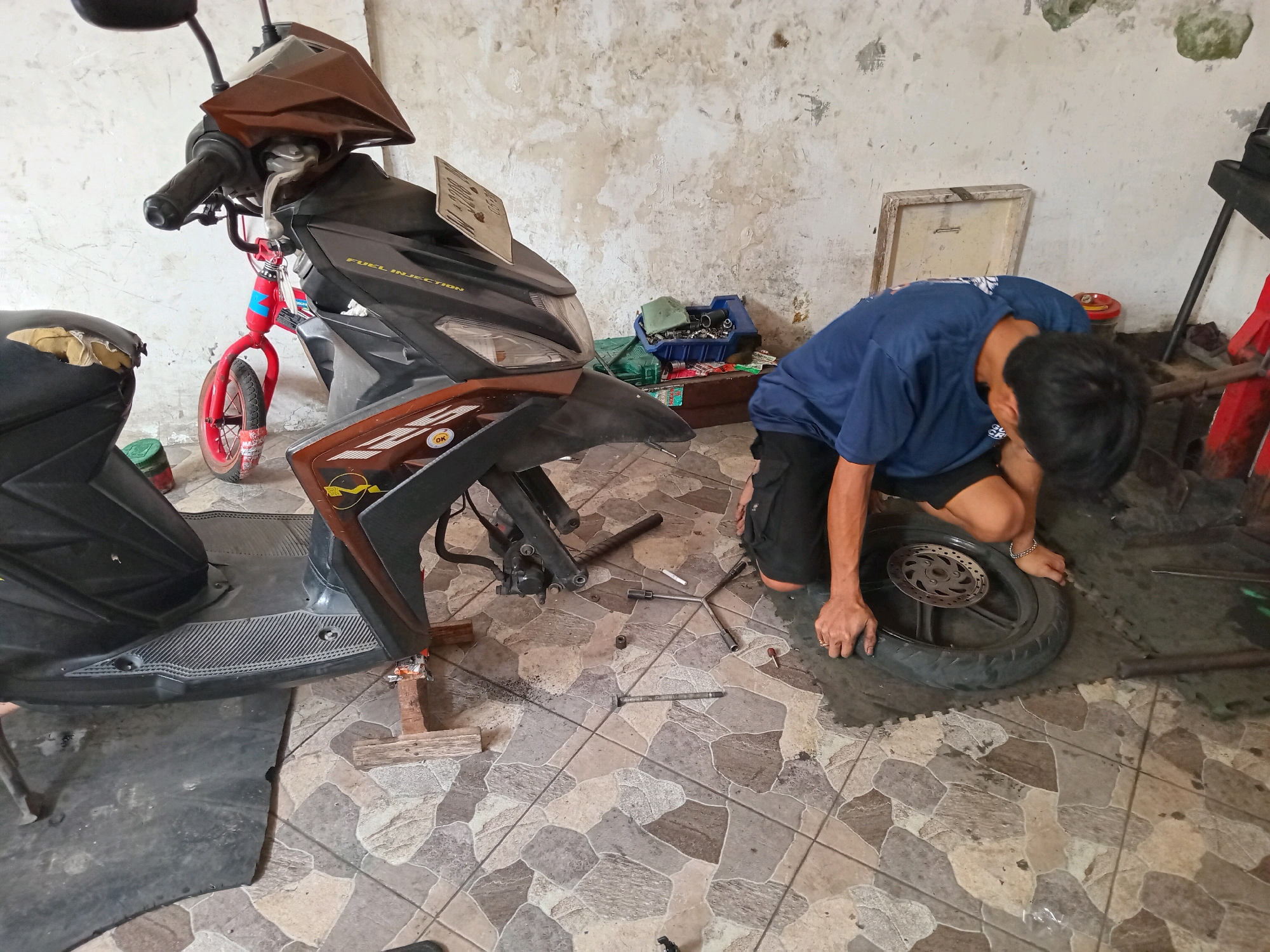 In Tebalo, Indonesia, a young male is seen repairing a motorcycle in what appears to be a workshop or garage. The motorcycle, a scooter-style model with black and brown fairings, is partially disassembled. The front wheel has been removed and is being worked on by the individual, who is kneeling on the floor. Tools, including a socket wrench, are scattered around the motorcycle. The environment is a tiled floor with a distressed, textured wall in the background. The scene suggests a typical repair or maintenance activity for a motorcycle.
