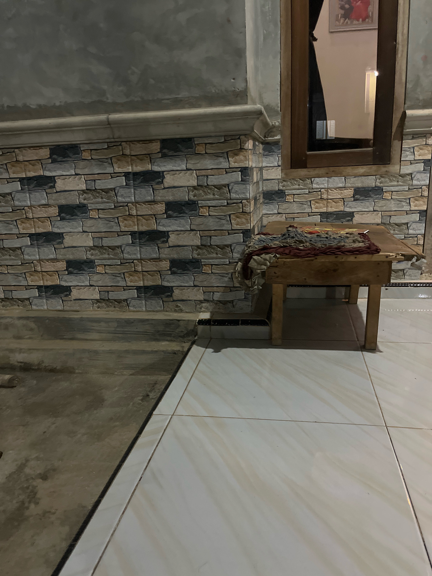 The image depicts an outdoor scene near a building in Kebumen, Indonesia. The foreground features light-colored tiled flooring, transitioning to a darker, rougher surface on the left. A small wooden table with four legs stands on the tiled floor, positioned against a wall covered in a stone-patterned tile. A piece of fabric, appearing to be a rug or textile, is draped over the table. The wall also has a decorative molding and a concrete section above. A window with a wooden frame is visible on the right side of the wall, reflecting the interior of the building which includes a framed picture. The lighting suggests it is either dusk or evening, or the area is dimly lit. There are no people or animals visible in the scene.