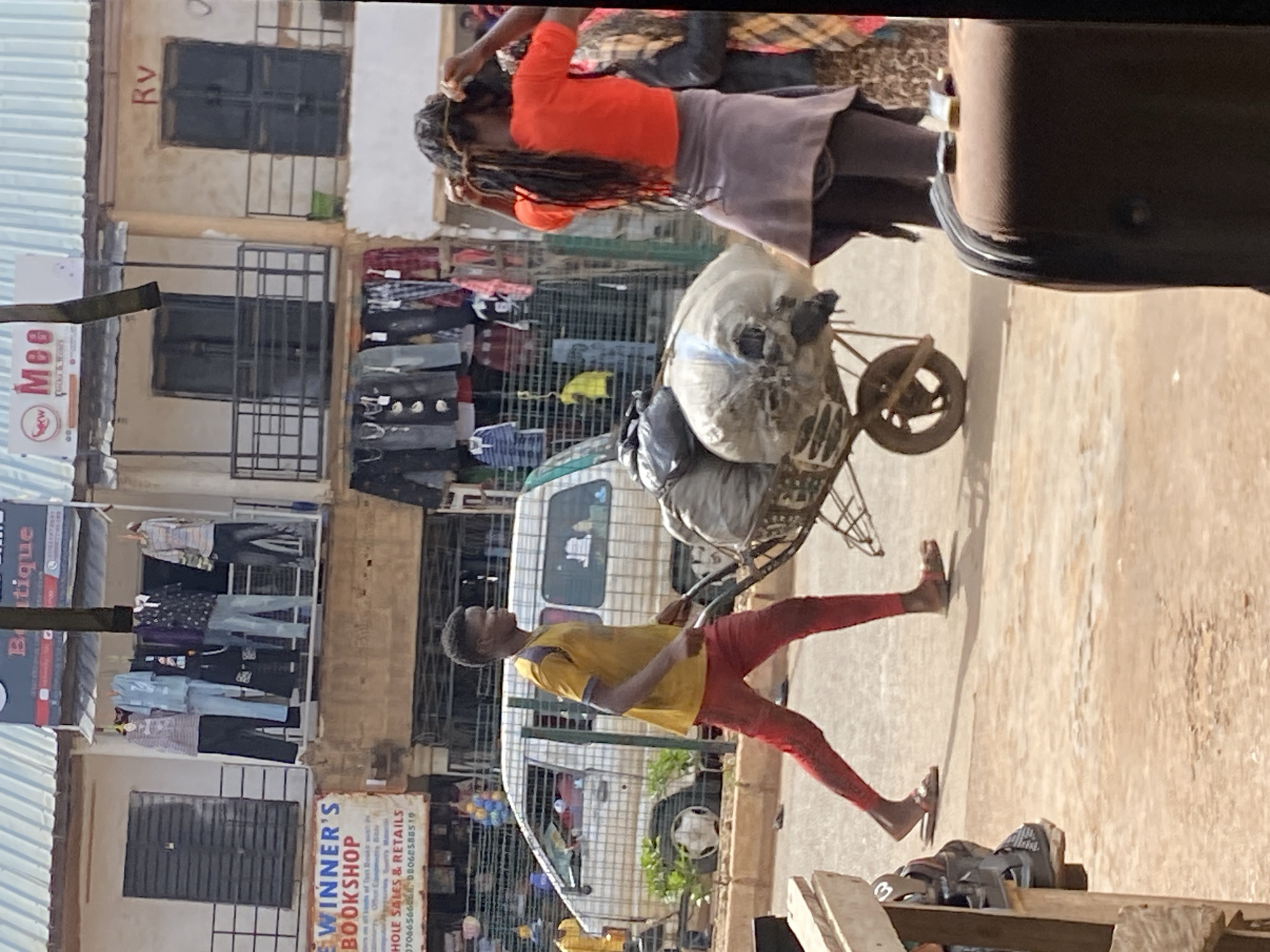 In Bukuru, Nigeria, a young man with dark hair and fair skin, wearing a yellow t-shirt and red pants, pushes a wheelbarrow loaded with large, white, burlap sacks. He is walking to the right. In the background, a white van is parked. A woman with dark, braided hair, wearing an orange shirt and a grey skirt, is tying her hair. She is standing to the right of the wheelbarrow. Buildings with corrugated metal roofs line the street. Clothes are displayed on racks outside some of the buildings. The ground is dirt and paved with asphalt. The scene appears to be a busy marketplace.
