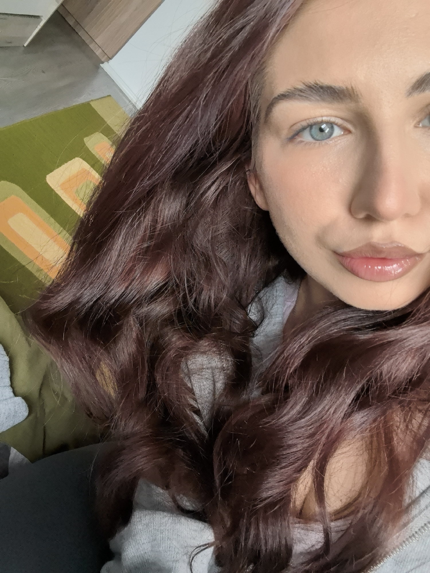 A selfie of a fair-skinned woman with long, wavy auburn hair and blue eyes. She is wearing a gray top. The background shows a glimpse of a green and orange patterned surface, along with a hint of a wooden surface. The location and city cannot be determined.