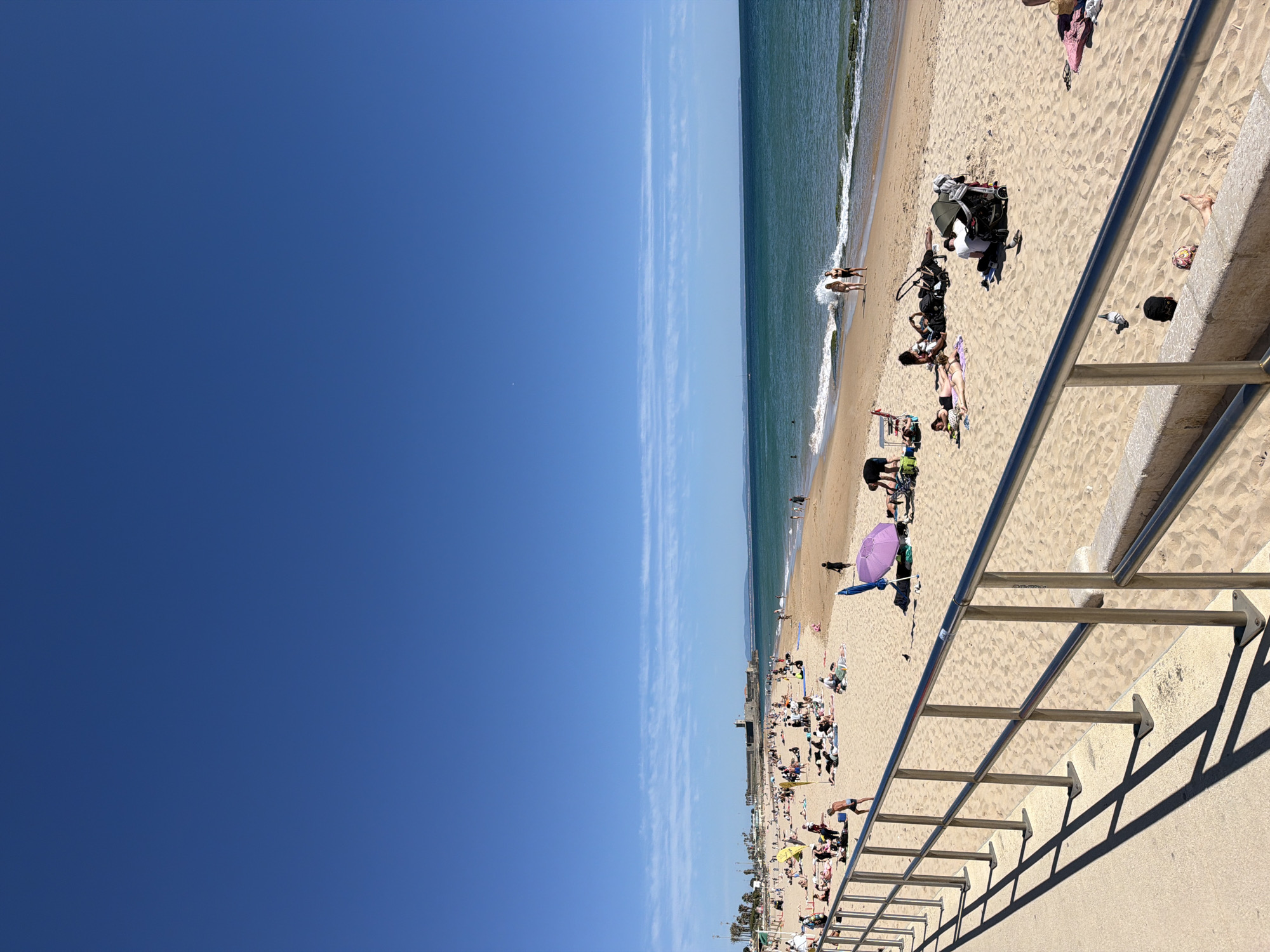 The image captures a sunny day at the beach. A clear, bright blue sky occupies the left half of the frame. On the right is the beach, lined with people enjoying the sun and surf. The ocean is a gradient of blue and green, with gentle waves meeting the sandy shore. In the foreground, a metal railing casts a shadow on a concrete surface. People are scattered across the beach, some sunbathing, others swimming in the sea, and a few walking along the shoreline. A purple parasol can be seen among the beachgoers. In the distance, a structure is visible near the beach, possibly indicating the location is in a developed area. Considering the city location in Cascais, Portugal, the beach could be one of the popular beaches in this coastal town.
