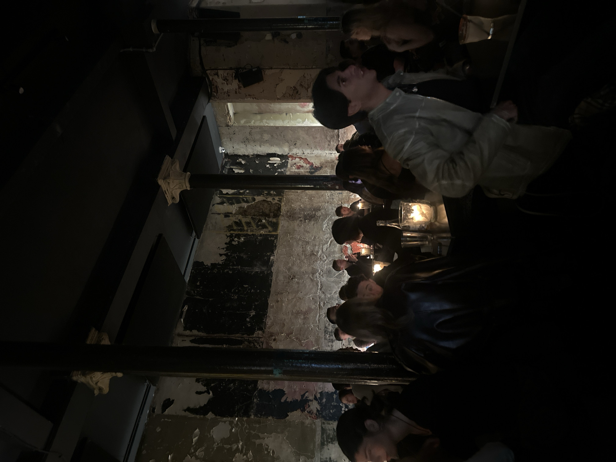 The image depicts a dimly lit interior space, possibly a bar or restaurant, in what appears to be an older building with exposed brick and peeling paint. A row of people are seated, likely at a counter or bar. The subjects seem to be engaged in conversation or observing something beyond the frame. There are candles on the counter.
The location cannot be determined with certainty from this image alone. However, the architectural style and atmosphere are consistent with the kinds of establishments that can be found in certain areas of London.