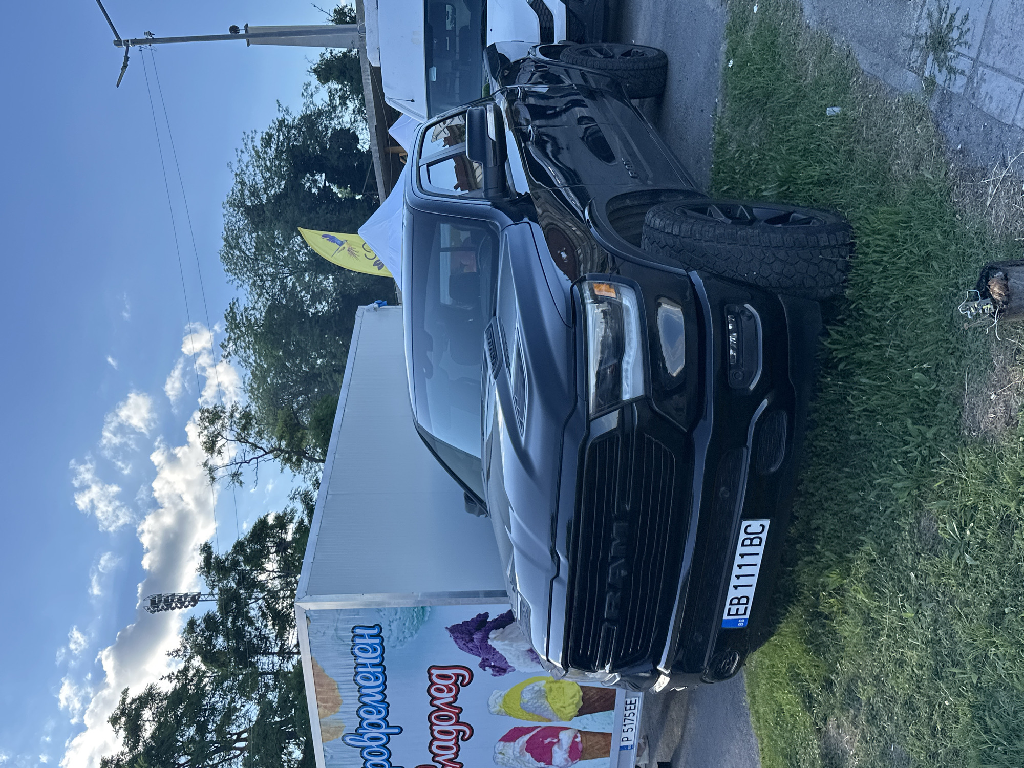 The image shows a black Ram 1500 pickup truck parked on a grassy area next to a white trailer with an advertisement for ice cream. The truck's license plate reads "EB 1111 BC". In the background, there are trees, a large stadium light, and a yellow flag with a bird logo. The scene appears to be an outdoor event or market, possibly in the city of Gorna Oryahovitsa, Bulgaria, given the Bulgarian license plate. The sky is blue with scattered clouds.