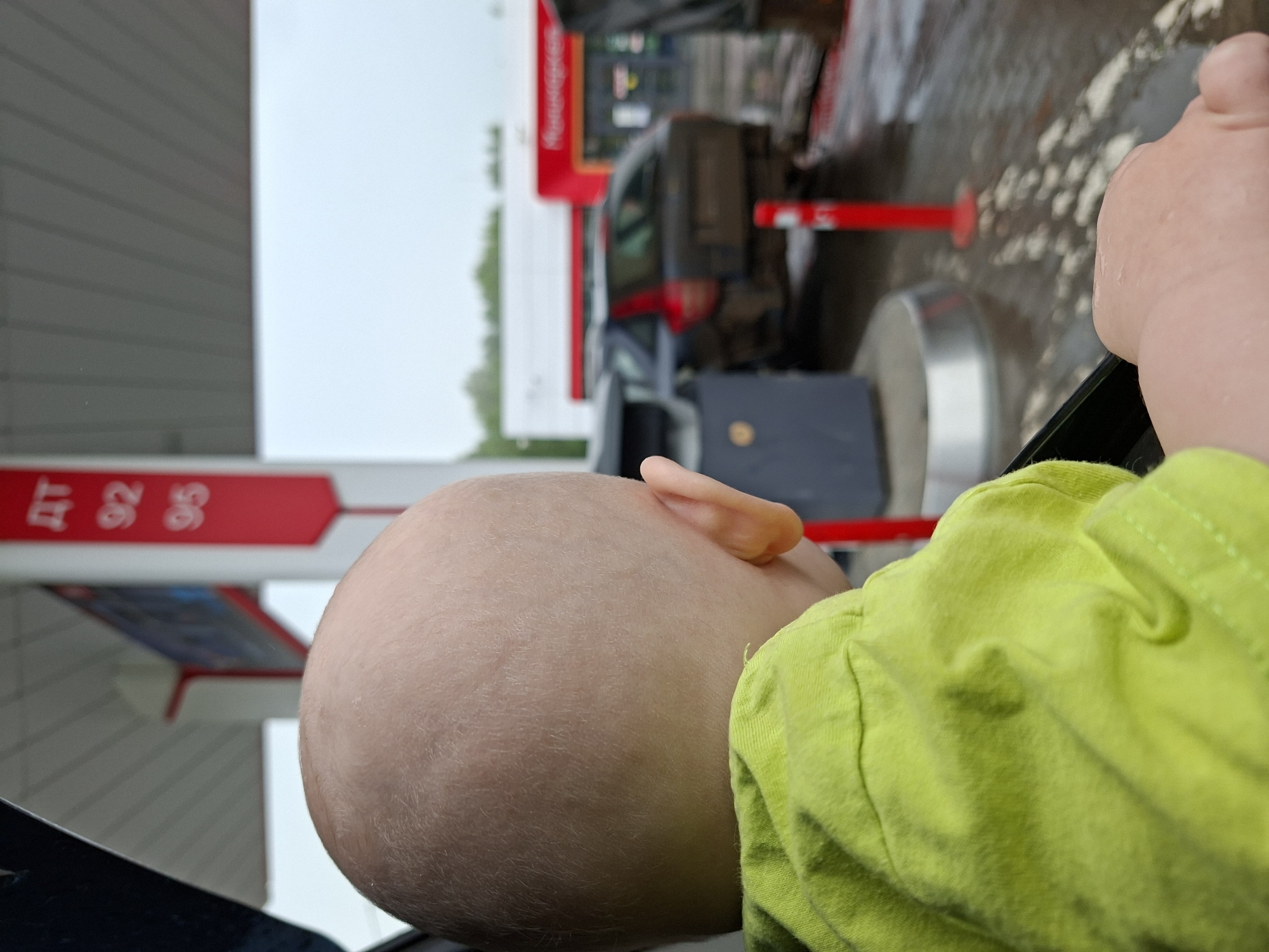 An infant's bald head, seen from behind, is in the foreground, facing towards a gas station under a cloudy sky. The infant wears a bright green t-shirt. In the middle ground, a dark-colored car is parked at a gas pump. The gas station canopy is visible, with a red sign displaying fuel prices: "ДТ 92 95". The ground is wet, indicating it has rained. The background shows buildings, possibly part of the gas station's convenience store, with red accents. The overall setting is a gas station in Krilovo, Russia.