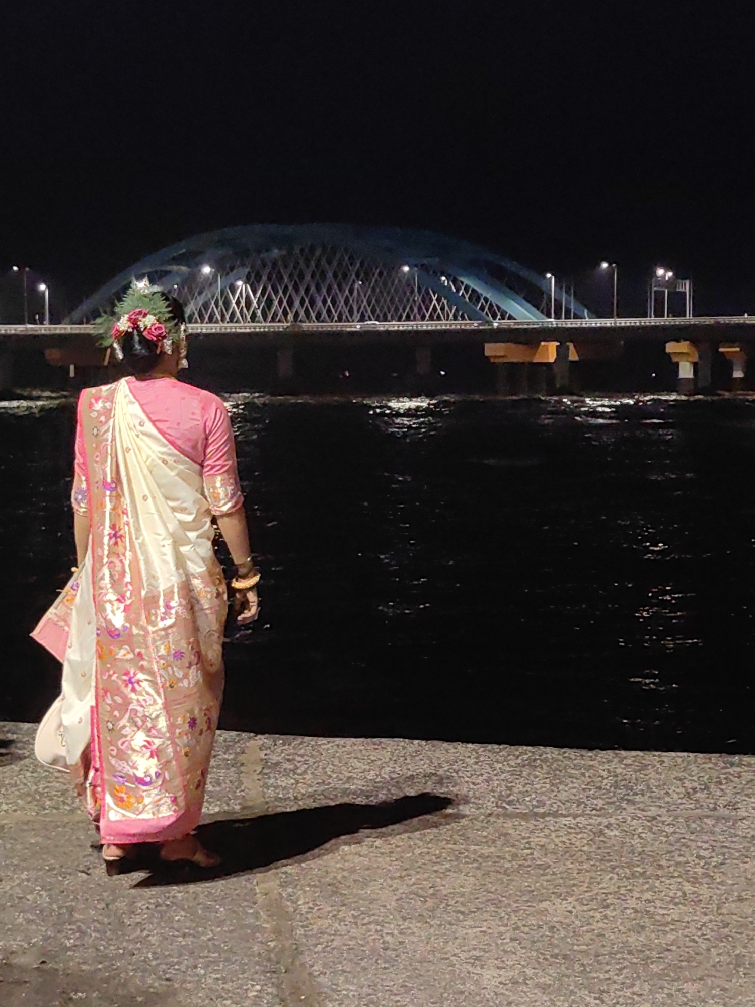 A person wearing a traditional Indian saree and adorned with flowers in their hair stands facing away from the camera, looking out at a body of water at night. Behind the person and the water is a large bridge with illuminated arches and a complex lattice structure. The water reflects the lights from the bridge and the surrounding city. The ground in the foreground is paved with textured material, and the person casts a long shadow. The scene appears to be a scenic urban waterfront at night. The location is likely the Bandra-Worli Sea Link in Mumbai, India, given the distinctive architecture of the bridge.