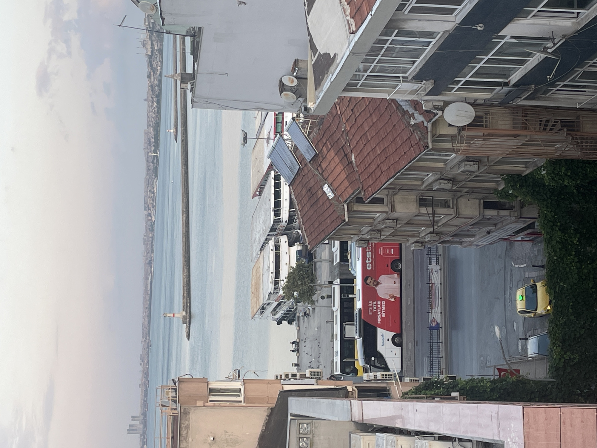 This is an eye-level, exterior, full shot taken from a residential building overlooking a waterfront area in Kadıköy, Turkey. The time of day appears to be late afternoon or early evening, with a diffused light and an overcast sky.
In the foreground, several multi-story buildings are visible, some with red tile roofs and others with white facades. Balconies and air conditioning units are common features on the buildings. A narrow, winding metal staircase is attached to one of the buildings. Lush green ivy climbs up the side of another building.
A street runs horizontally across the mid-ground, with a yellow taxi visible parked on the right side of the street, illuminated by its green taxi light. A dark-colored car is parked behind the taxi.
Beyond the street, a paved promenade area is populated by a flock of pigeons and two silhouetted figures sitting on a bench. A large, red and white tour bus with an advertisement featuring a smiling man and text in Turkish is parked in front of the promenade. Another yellow and black bus is parked next to it.
Further back, several large, multi-deck ferries are docked along the waterfront. In the distance, a lighthouse stands on a pier extending into the water. The background features a hazy cityscape with buildings and faint skyscrapers, indicative of a large urban area. The sea is calm, with a light blue hue.