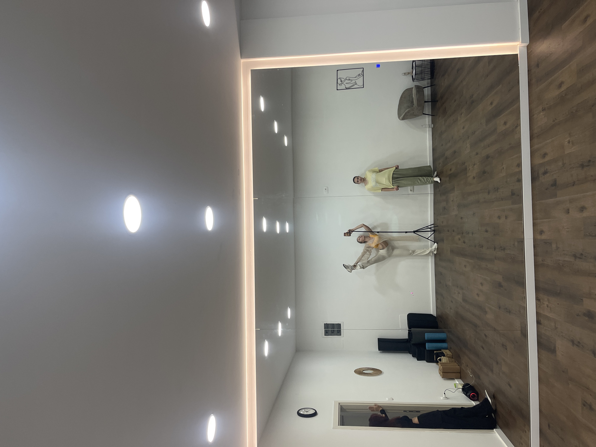 The image depicts an indoor studio or gym in Budva, Montenegro, characterized by white walls and dark wood-look laminate flooring. The ceiling is illuminated by multiple recessed circular lights. A large, floor-to-ceiling mirror occupies the central wall, framed by a luminous strip, reflecting the room and its occupants.
Three individuals are present. On the far left, a person with dark hair, dressed in dark clothing, is partially visible in a doorway, leaning against the frame with their right hand raised. In the reflection within the large mirror, two women are visible. The woman on the left (viewer's right in the mirror) is fair-skinned, wearing a yellow crop top, light-colored pants, and white athletic shoes. She is performing a high leg extension, holding her raised right foot with her right hand, while holding a smartphone in her left hand, possibly for recording or a selfie. A black tripod stands beside her. To her right (viewer's left in the mirror), a second fair-skinned woman stands upright and relaxed, wearing a light yellow t-shirt and green wide-leg pants.
The environment contains various objects: a black circular wall clock near the doorway, a circular woven decorative piece on the wall further right, and a stack of black yoga mats and blue yoga rollers on the floor along the left wall. A black portable speaker with red lighting is on the floor near the mats. On the right side of the mirrored wall, a gray upholstered armchair and a small black wire side table are present. Above the armchair, a framed minimalist line art drawing of a figure is mounted on the wall. The mirror reflects the ceiling lights and portions of the opposite wall.