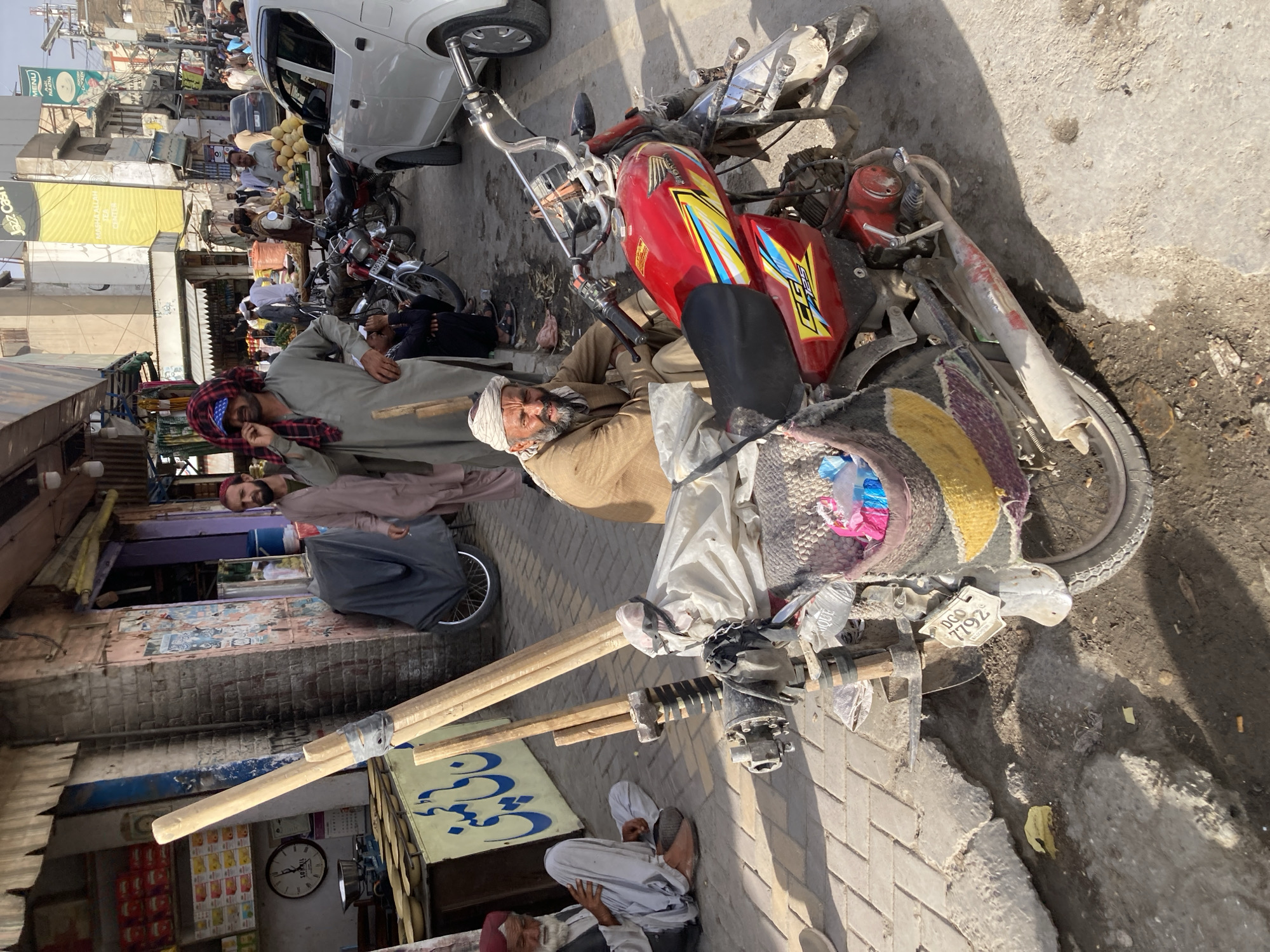 The image depicts a street scene in Quetta City Tehsil, Pakistan. The foreground is dominated by a heavily laden motorcycle parked on a brick-paved sidewalk. An older man wearing a traditional brown outfit and turban is partially reclining on the motorcycle. The motorcycle has a red tank with colorful decals. Behind him, a man in a grey shirt and plaid head covering stands. Another man behind the pair is wearing a mauve shirt and a cap. In the background, a street with various shops, vehicles, and pedestrians can be seen. A white car is parked nearby. Several men are sitting and standing among bags of what appears to be melons. The buildings are a mix of brick and painted structures. A sign with what appears to be an Arabic script is visible on a storefront. The lighting suggests it is daytime. Wooden poles are strapped to the front of the motorcycle. The license plate is visible and says "DCQ 7792."