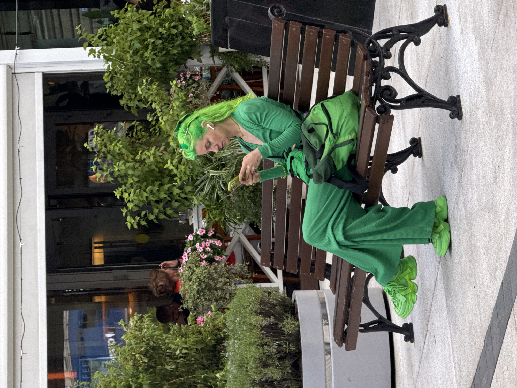 A woman with bright green hair is sitting on a park bench, looking at her phone. She is wearing a matching green outfit, including pants, a long-sleeved shirt, and chunky sneakers. A green backpack is on the bench next to her. In the background, there are trees, flowers, and people sitting at tables. The scene appears to be outdoors, possibly in a cafe or restaurant with outdoor seating. The woman is the central focus of the image, with her vibrant hair and clothing standing out against the more subdued background. The overall impression is one of a casual, urban setting.