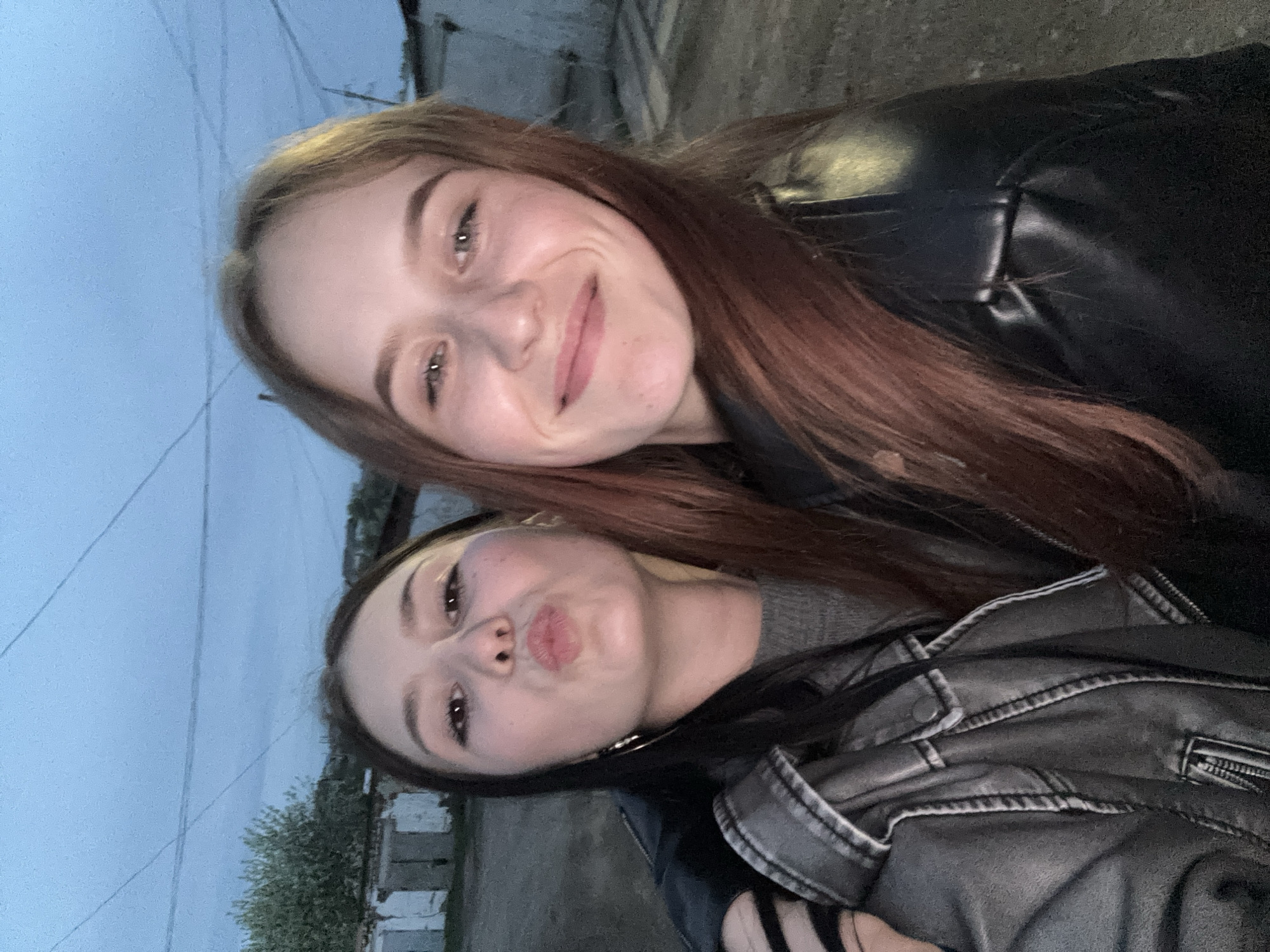 Two young women are posing for a selfie outdoors. The woman at the top of the frame has reddish-brown hair and is smiling. She is wearing a black leather jacket. The woman at the bottom of the frame is making a kissing face. She has dark hair and is wearing a gray sweater and a gray leather jacket. The background appears to be an urban environment, potentially in the city of Фоки, Russia, given the prompt's request. There are indistinct buildings and trees visible in the background. The lighting suggests it is dusk or a cloudy day.