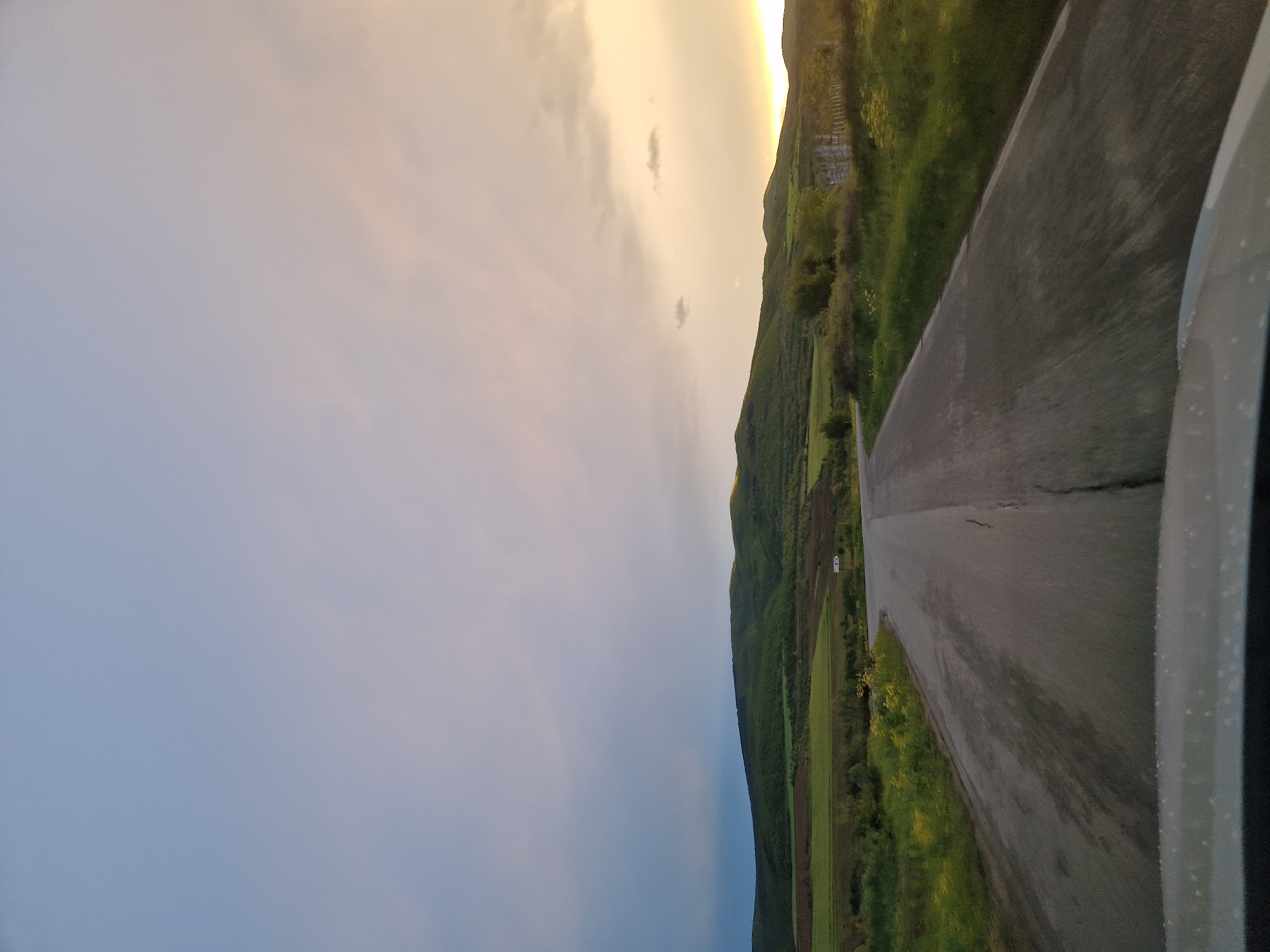 The image captures a serene rural landscape, likely near Bata, Bulgaria, from the perspective of a moving vehicle during either sunrise or sunset. A paved, two-lane road stretches diagonally from the bottom right foreground towards the upper left, showing some minor cracks on its surface. On the far right, a portion of a white or light gray vehicle body is visible, adorned with small water droplets, indicating recent rain or heavy dew.
To the right of the road, a verdant, undulating hillside or mountain range rises, extending into the distance under the warm, golden light of the low sun. On the left side of the road, expansive agricultural fields, a mix of vibrant green crops and darker tilled earth, spread towards the base of the distant hills. A small white rectangular object, possibly a sign or a distant structure, can be seen in the fields to the left of the road.
The sky, occupying a significant portion of the upper frame, transitions from a muted blue-gray in the middle-left to a brilliant yellow and orange glow on the far right horizon, illuminated by the setting or rising sun. Scattered, wispy clouds are visible against the luminous sky. No people are visible, and the scene conveys a sense of peaceful travel through a picturesque natural environment.