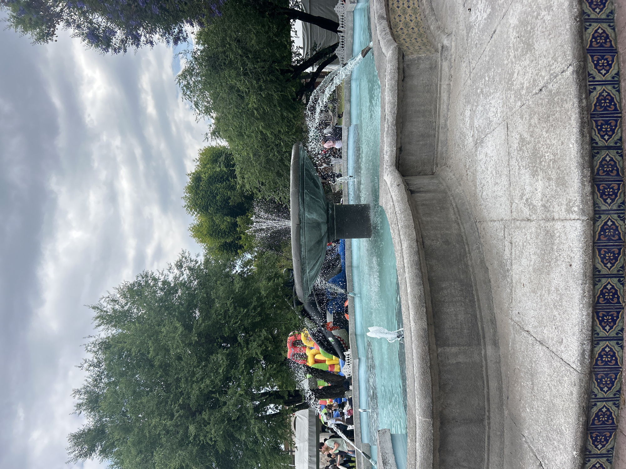 The image shows a fountain in an outdoor setting, possibly a park or plaza. The fountain is constructed of concrete and features blue and white patterned tiles along its edge. Water is actively flowing from the fountain's central structure. In the background, trees with green foliage are visible. There are numerous people in the distance and an inflated playground structure, adding to the atmosphere of a public gathering space. The sky is overcast with grey clouds.