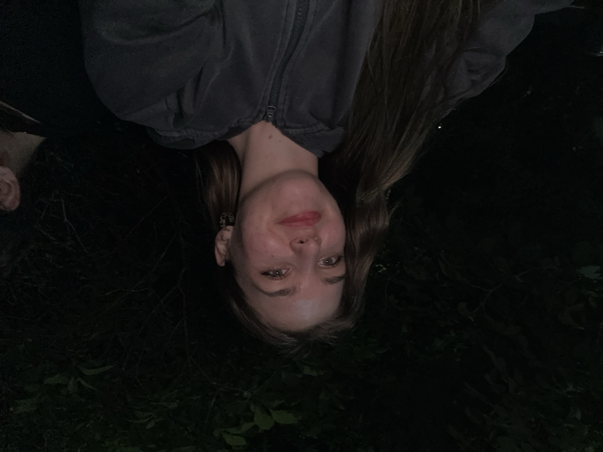An upside-down, low-angle photograph shows a woman with fair skin, wearing a gray zippered jacket, with long brown hair. Her face is centered in the frame, with her eyes looking upwards towards the camera. She has red lipstick. The background is dark, suggesting nighttime. Green foliage is visible towards the bottom of the frame. The location is not specified.