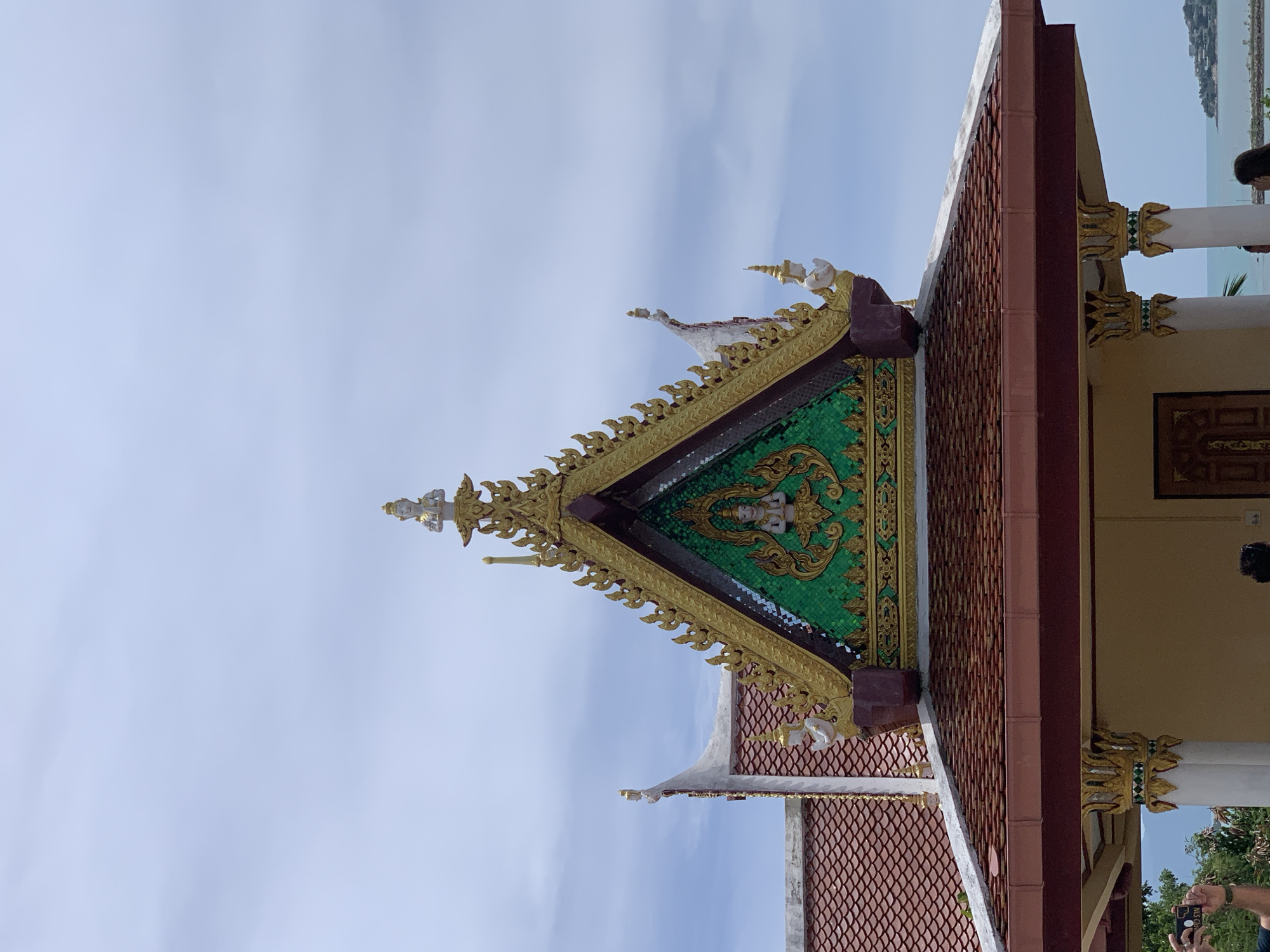 This is a medium-shot, eye-level, vertical-orientation photograph of the ornate roof of a building, likely a temple or shrine, located in the Ko Samui District of Thailand. The building's roof is tiled with reddish-brown, curved tiles. The main focus is the highly decorative gable of the roof, which is triangular in shape. This gable is adorned with intricate gold-colored ornamental carvings that resemble flames or elaborate patterns. In the center of the gable, against a backdrop of green mosaic tiles, is a white statue of a person in a prayerful or respectful pose, with hands pressed together. Above this statue, at the very peak of the gable, is another smaller white statue of a person with a crown. The roof's edges are also decorated with gold-colored patterns, and small white figures in similar poses are positioned on the corners of the roof's lower edges. To the left of the main gable, part of another roof section is visible, also covered in reddish-brown tiles and topped with a white, pointed decorative element. The sky above is a pale blue with scattered white clouds. The lower portion of the image shows the building's exterior walls, which are a light beige color, and two white columns with gold and green decorative caps. A sliver of the ocean and a distant coastline with buildings can be seen to the right, indicating a coastal location. In the lower left corner, the hands of a person holding a smartphone are visible, as if taking a photograph of the scene. Another person's head and shoulders are visible near the bottom center of the image.