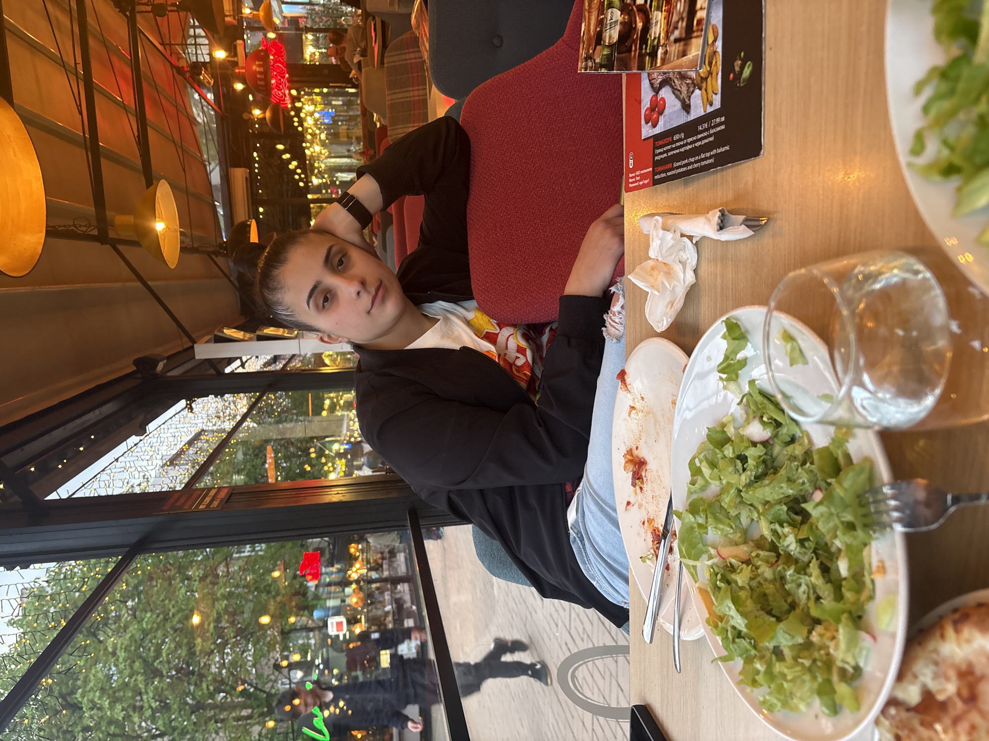 This is an eye-level, horizontal photograph of a young individual seated at a wooden dining table in what appears to be a restaurant or cafe in Sofia, Bulgaria, during the evening. The person, with dark hair tied back, wears a black jacket over a white and colorful t-shirt, and light blue ripped jeans. They gaze directly at the camera with a neutral to slightly serious expression. Their left arm is casually placed behind their head, and their right arm rests on the table.
The dining table in front of the person holds plates with remnants of food, including a green salad with radishes, and another dish with a red sauce. An empty drinking glass, a fork, and a knife are also visible. A menu stands upright on the table, displaying details for "TOMAHAWK 650 g/d" priced at "27.99 лв" (Bulgarian Lev), described as a "Grand pork chop on a flat top with balsamic reduction, roasted potatoes and cherry tomatoes" in both Bulgarian and English. Other visible text includes "WID" and "June."
The background showcases the warm ambiance of the establishment, with several suspended yellow-toned light fixtures and other dining furniture. Through a large glass window or partition to the left, an outdoor urban scene is visible with green trees and numerous string lights, suggesting a lively evening atmosphere in Sofia. A red "Coca-Cola" sign is also faintly visible in the background. The overall scene suggests a relaxed dining experience.