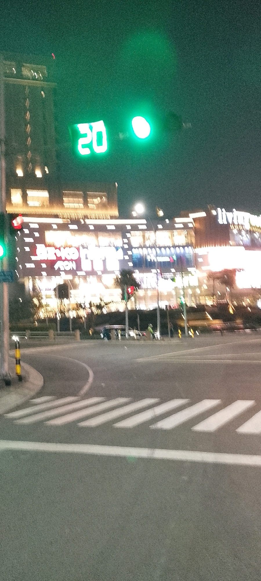 The image depicts a nighttime street scene in Gunung Putri, Indonesia, likely near a commercial area. A digital traffic signal displays the number "20", indicating a countdown timer. The background features a brightly lit building with illuminated windows and signage, possibly a shopping center or office complex. The street itself is asphalt, with a visible crosswalk. Several vehicles, including cars and what appears to be a motorcycle, are present on the road. Pedestrians can be seen in the background, one of whom is wearing a light-colored jacket. The overall atmosphere is urban and nocturnal, with artificial lighting dominating the scene.