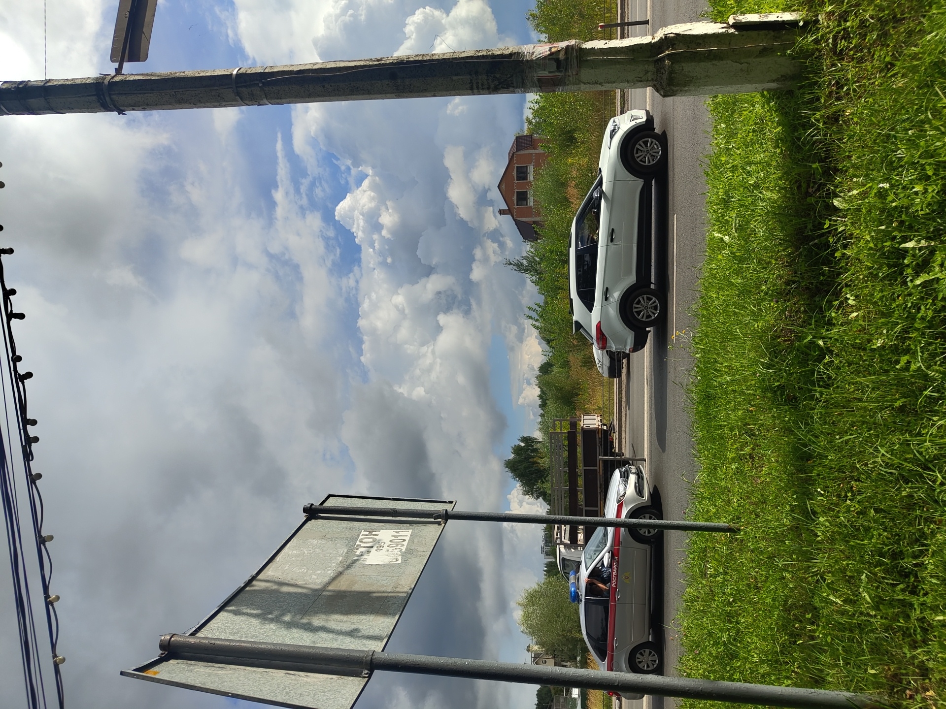In Dergayevo, Russia, a street scene unfolds under a partially cloudy sky. A road runs horizontally across the image, bordered by lush green grass. On the road, a white SUV is visible, and to its left, a silver sedan with a blue light on its roof and the word "РОСГВАРДИЯ" (Rosgvardiya) printed on its side is stopped. The driver of the silver sedan is partially visible. Behind the sedan, a flatbed truck is parked, and further down the road, another vehicle is partially visible. A tall, weathered concrete utility pole stands prominently on the right side of the frame, partially obscuring a section of the sky. In the background, dense green foliage lines the road, with a red-brick house with a dark roof situated behind the trees. A large, gray metal signpost stands on the left side of the image, with a visible sign on it bearing Cyrillic text and numbers. Overhead, electrical wires with small lights are strung across the top of the frame.