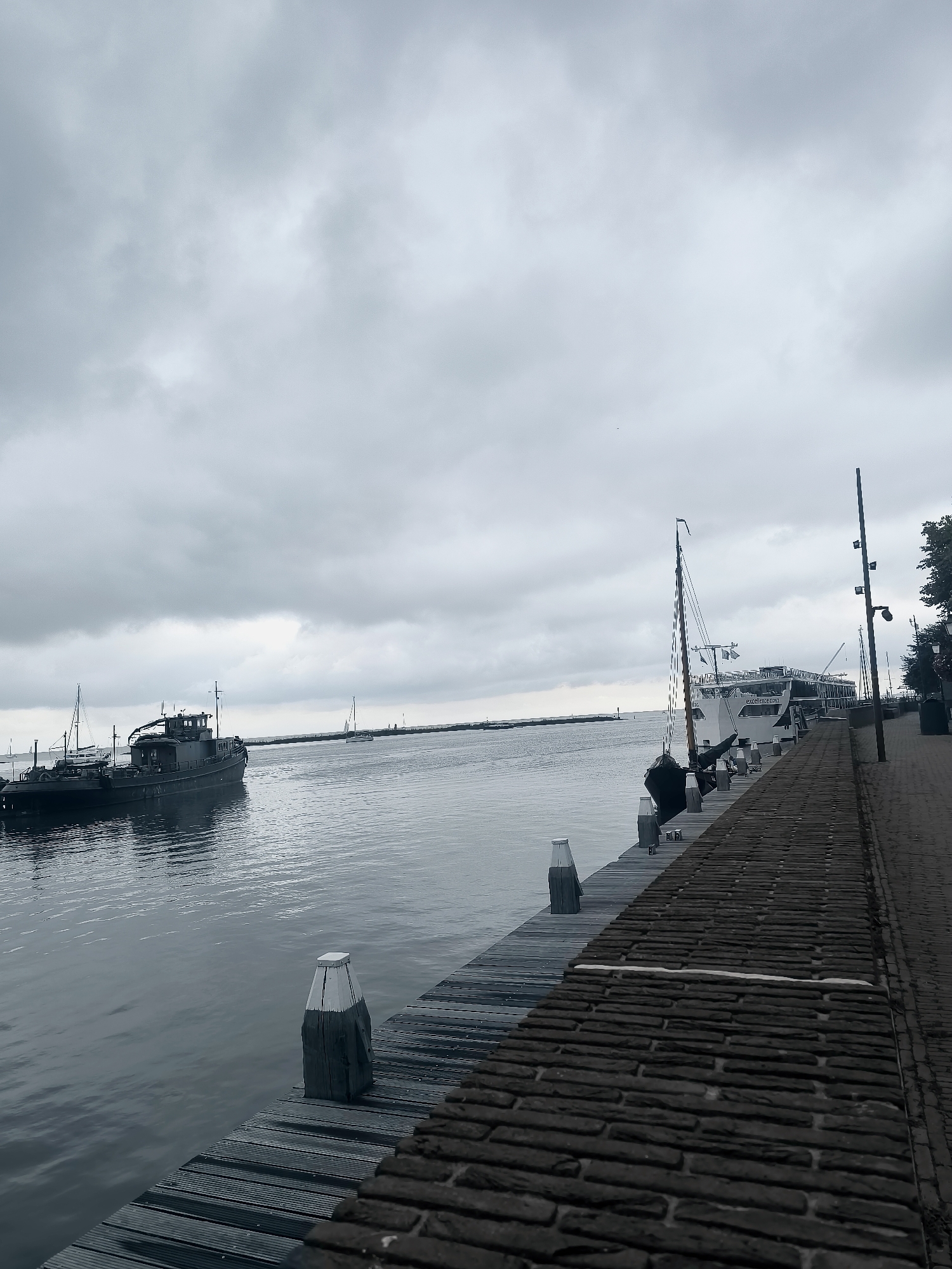 The image depicts a cloudy day at a harbor in Hoorn, Netherlands. In the foreground, a wooden pier runs along the right side of the frame, paved with dark bricks. The pier has several bollards. To the left of the pier, a calm body of water reflects the overcast sky. A dark-colored tugboat is visible in the water to the left, with other boats, including a sailboat, further in the distance. A large white boat is docked at the pier on the right side of the image. The atmosphere is somber due to the cloudy conditions.