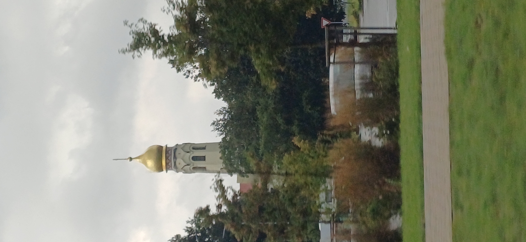 A close-up, vertical shot captures a gold-domed bell tower peeking through lush green trees against a cloudy sky. The bell tower has a classic architectural design with multiple levels and arched windows. The trees are a mix of deciduous and evergreen, with some showing autumnal colors. In the foreground, a patch of manicured green grass is visible, separated from the trees by a paved pathway. The scene appears serene, with no people or active subjects present. The overall impression is one of a peaceful, natural setting within an urban environment.