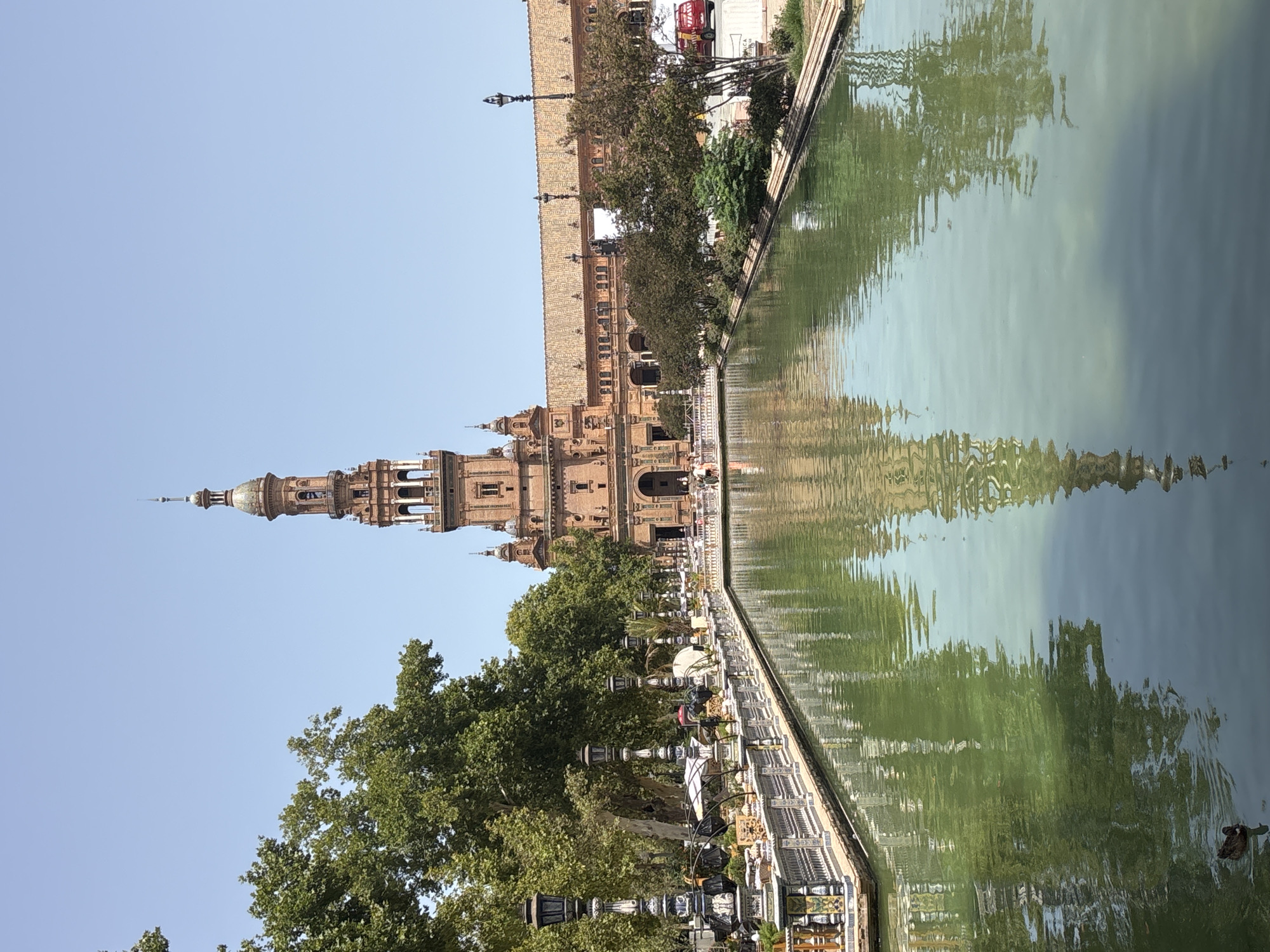 The image depicts the Plaza de España in Seville, Spain, on a clear, sunny day. A wide canal, reflecting the surrounding architecture and trees, dominates the foreground. A lone duck swims in the green water on the left side of the canal. Along the edge of the canal, a decorative tiled railing is visible, with ornate lampposts and some outdoor seating arrangements behind it. In the background, a large, ornate brick building with a prominent tower stands under a clear blue sky. Trees with lush green foliage line the canal and partially obscure the building. The architecture suggests a historic and grand European setting.