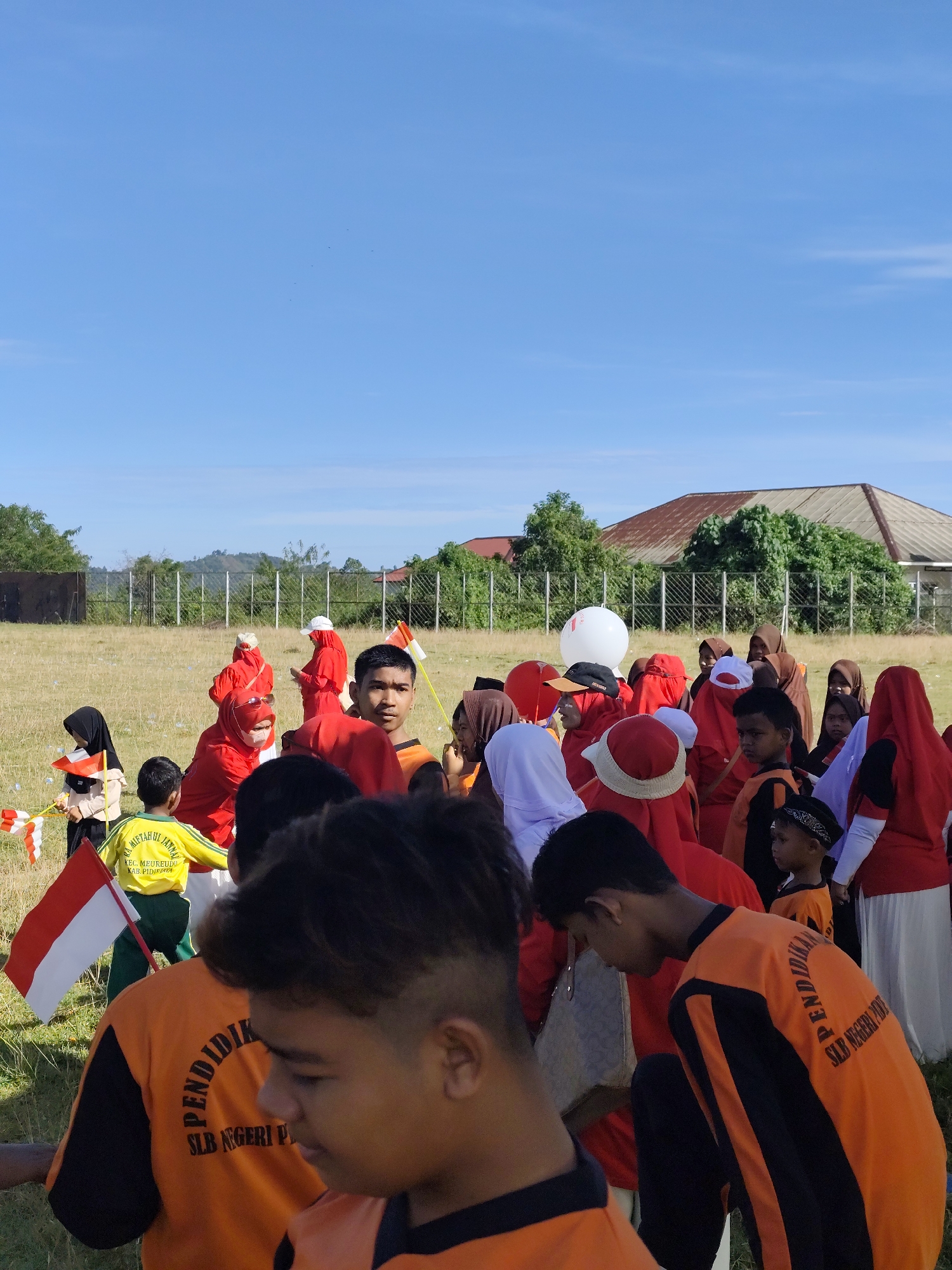 A group of people, primarily children, are gathered outdoors in a grassy field under a clear blue sky. Many are wearing red clothing and head coverings, while others are in orange shirts. Several children are holding small Indonesian flags. In the background, there is a fence, trees, and a building with a brown roof. The overall scene appears to be an outdoor event or gathering.