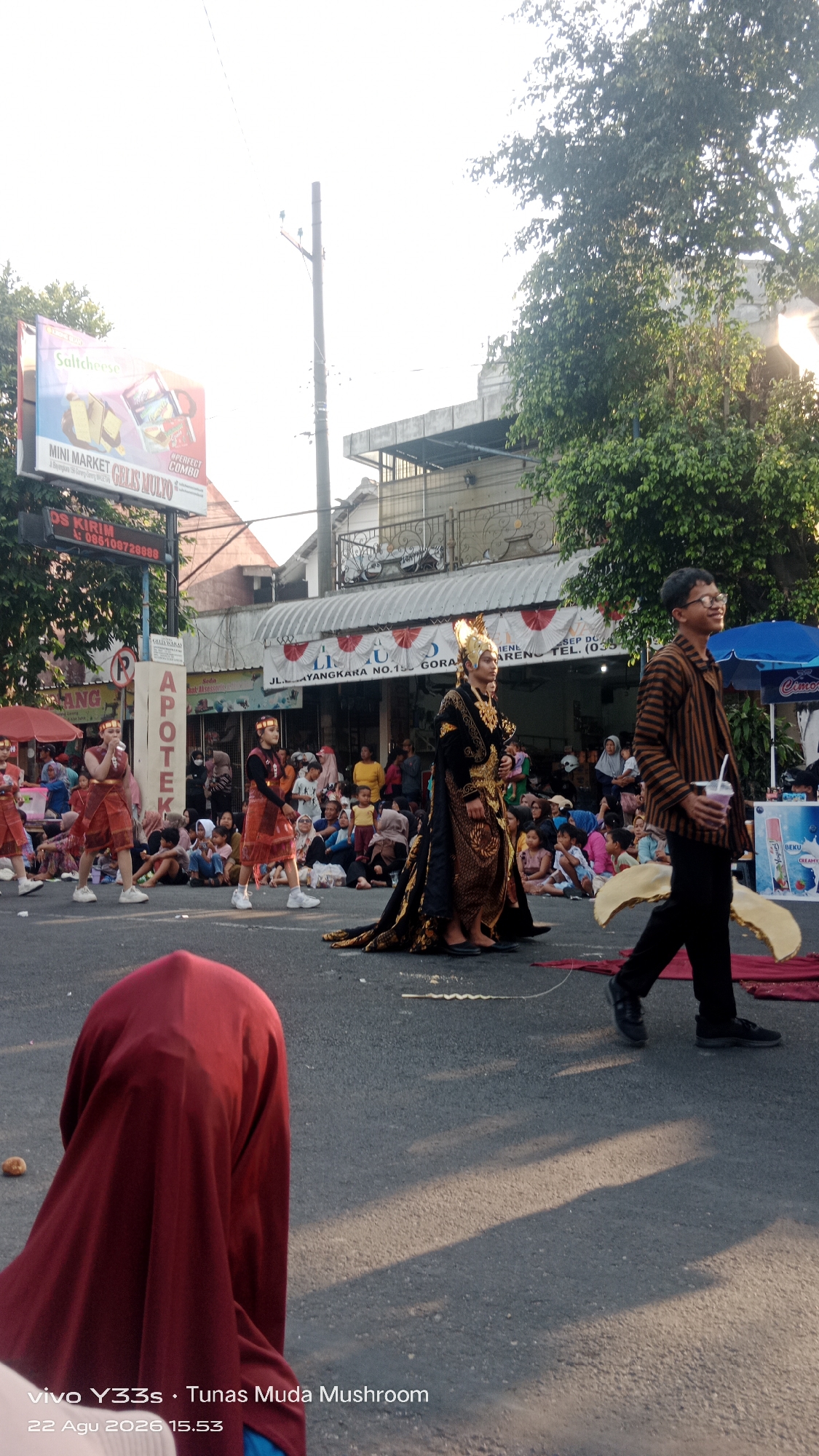 The image depicts a street scene in Genengan, Indonesia, during what appears to be a cultural event or performance. In the foreground, a person wearing a red hijab is partially visible, facing away from the camera. The middle ground features a procession or performance with individuals in traditional attire. Two individuals in ornate, gold-trimmed black robes with elaborate headdresses stand out, one walking towards the right of the frame and another with his back to the camera. Several children and young people in similar red and gold costumes are positioned to the left, seemingly part of the performance. A crowd of spectators is seated on the ground and standing along the street, observing the event.
In the background, buildings line the street, with storefronts and awnings visible. A billboard for "MINI MARKET GELIS MULYO" is prominently displayed on the left. A utility pole stands near the billboard. Trees with dense foliage are present on the right side of the image, casting shadows on the street. The lighting suggests it is late afternoon, with the sun casting long shadows. The overall atmosphere is one of a community gathering celebrating local culture.