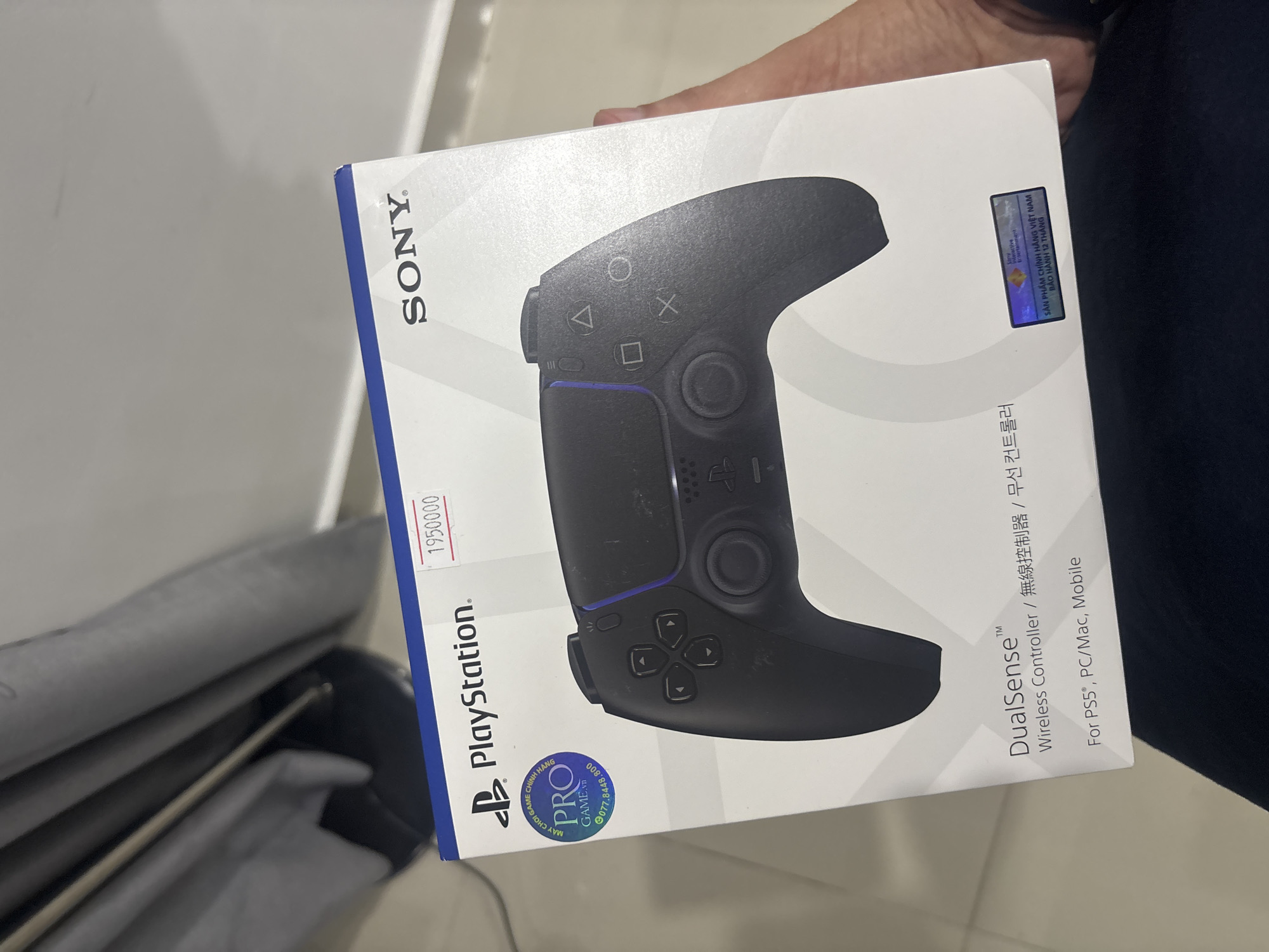 The image shows a black PlayStation 5 DualSense wireless controller box. The box is white with blue accents. The PlayStation logo and the word "PlayStation" are printed on the front of the box. The Sony logo is printed in the upper right corner. A sticker with a price of "195000" is present on the top right of the box. There is also a circular sticker with "PRO GAME.vn" and a phone number on the left side of the box. A holographic sticker indicating "SẢN PHẨM CHÍNH HÃNG VIỆT NAM BẢO HÀNH 12 THÁNG" is on the bottom right. The controller itself is partially visible on the box, showing the buttons and handles. The image appears to be taken indoors, with some curtains visible in the background. A person's hand is holding the box.